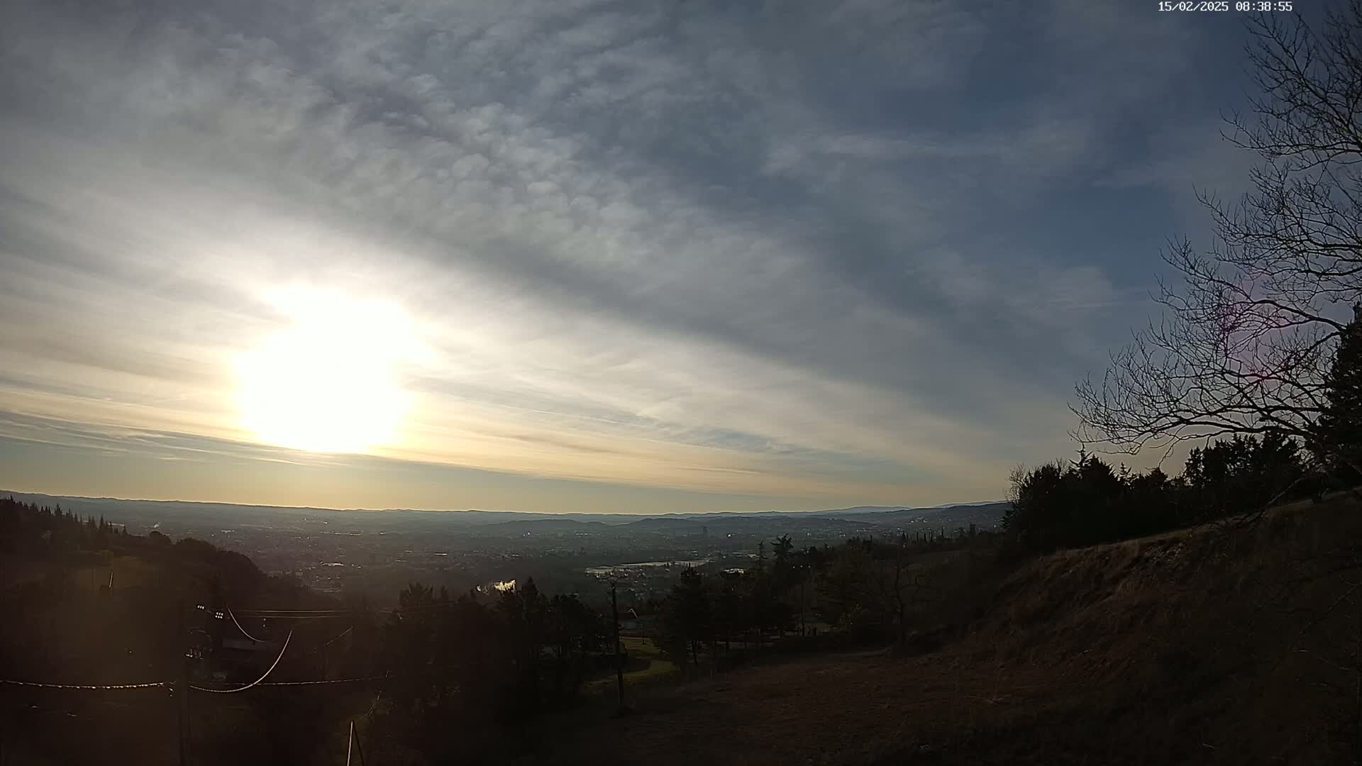 A partly cloudy sunset over a distant city viewed from a hilltop, with bare trees silhouetted against the sky.