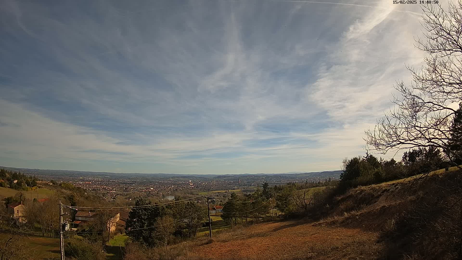 A sprawling, distant city is visible beneath a mostly clear sky with thin, wispy clouds, viewed from a hillside with sparsely wooded areas and several houses.