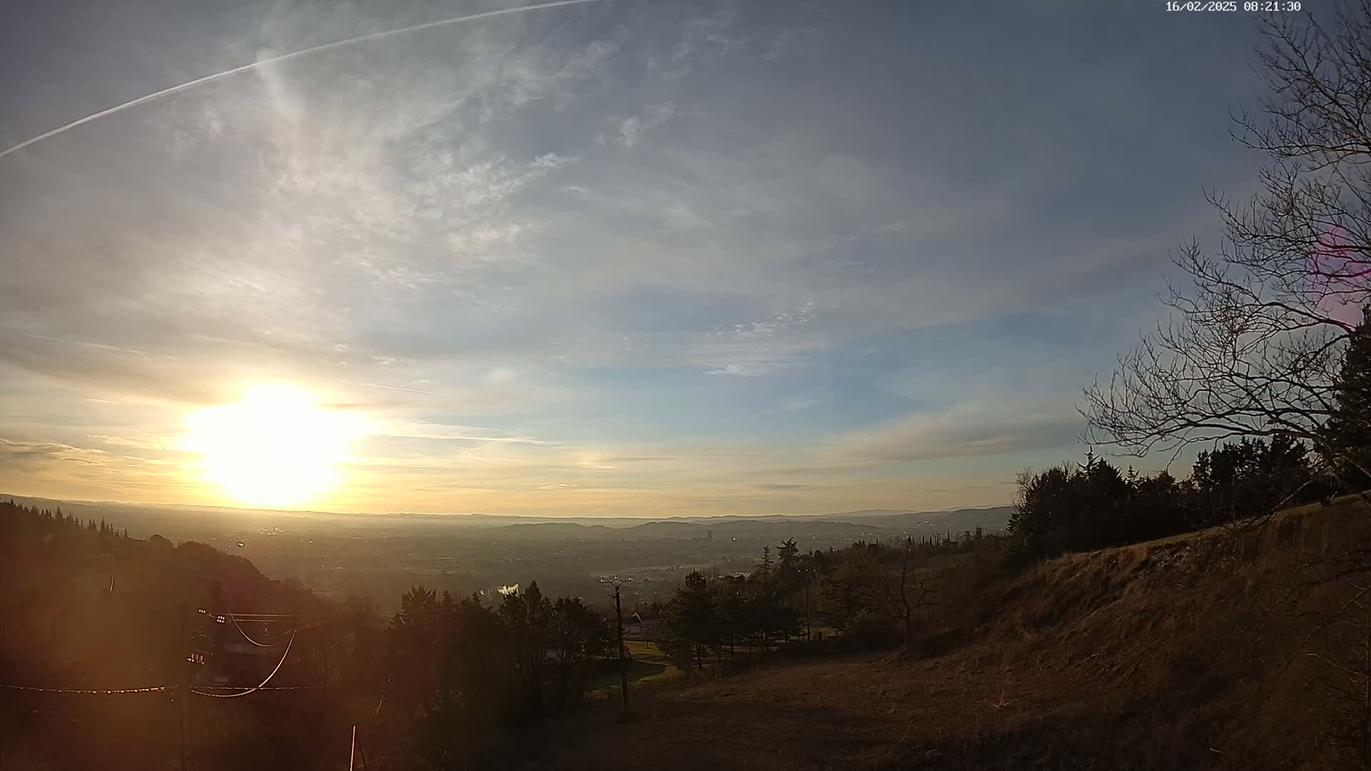 A bright sun sets over a hazy valley, partially obscured by a thin layer of clouds, with dark, leafless trees silhouetted against a hillside in the foreground.
