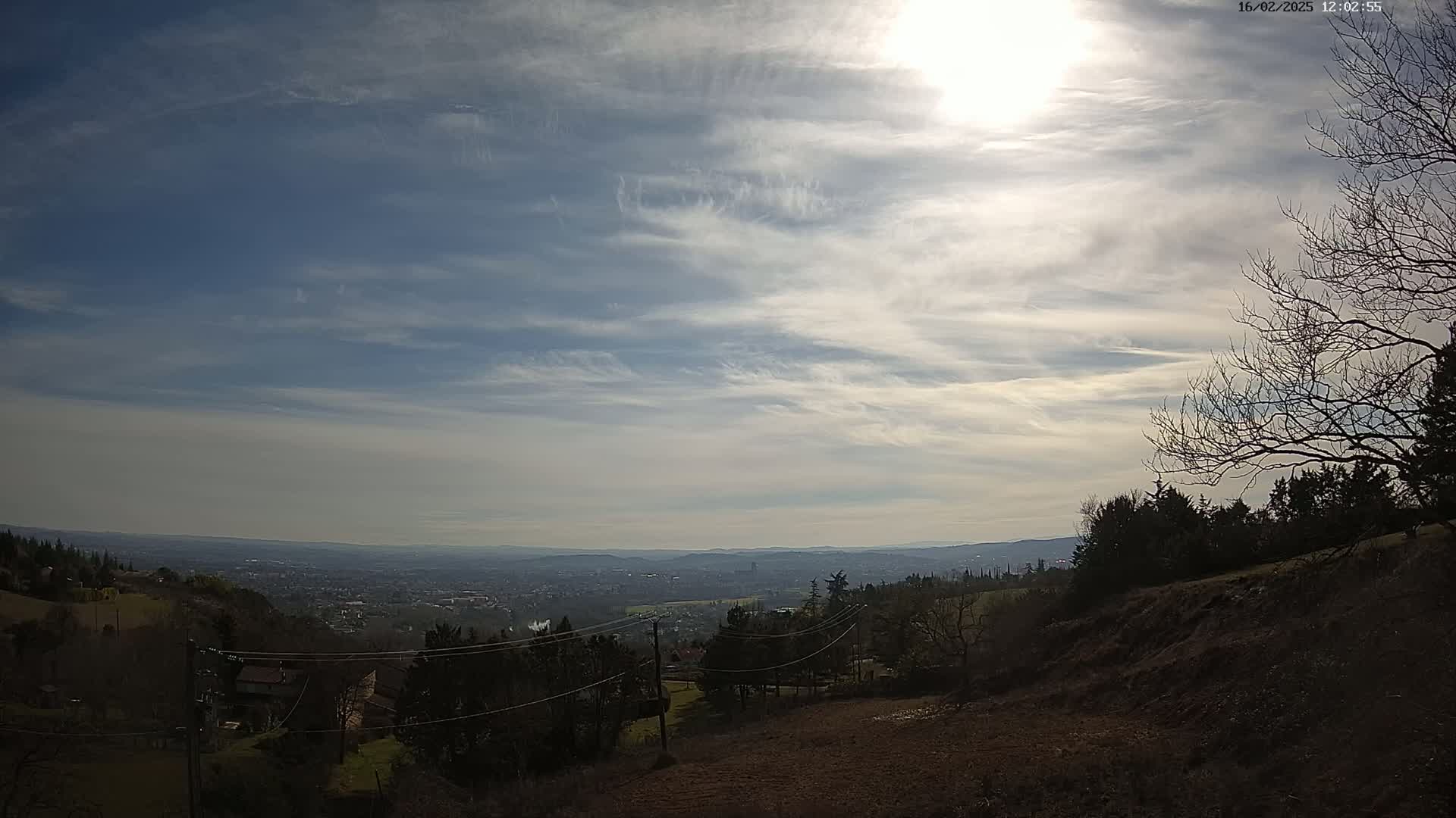 A view from a hillside shows a partly sunny sky with thin clouds over a distant city and rolling hills.