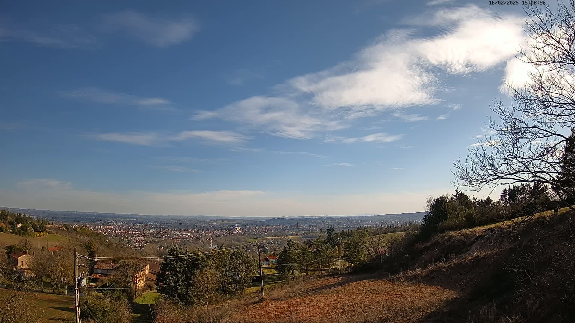 A mostly sunny sky with scattered clouds overlooks a hilly landscape featuring a distant city and several houses nestled amongst leafless trees.