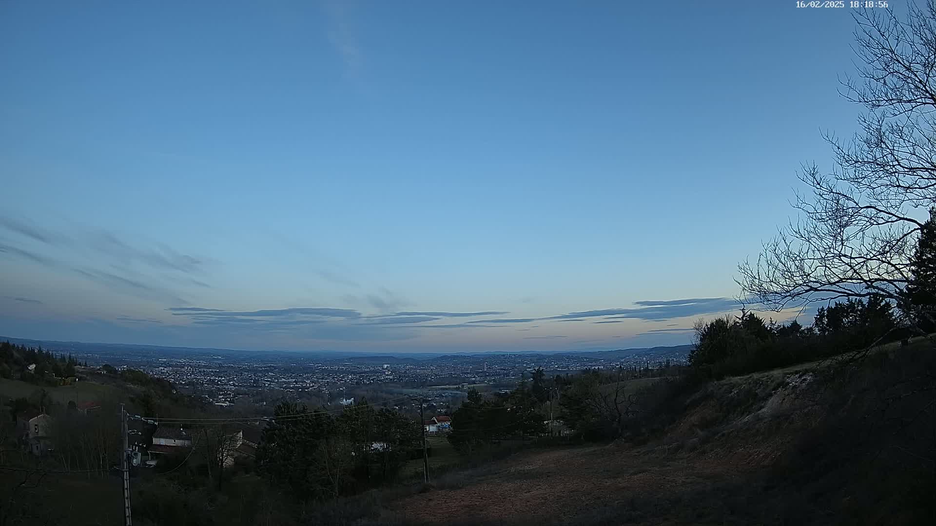A view from a hilltop at dusk shows a sprawling city in the distance under a mostly clear, pale blue sky with a few thin clouds.