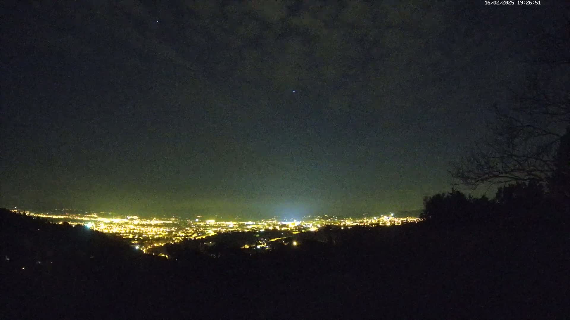 A nighttime view from a hilltop shows a city's lights spread out below under a mostly clear, dark sky.