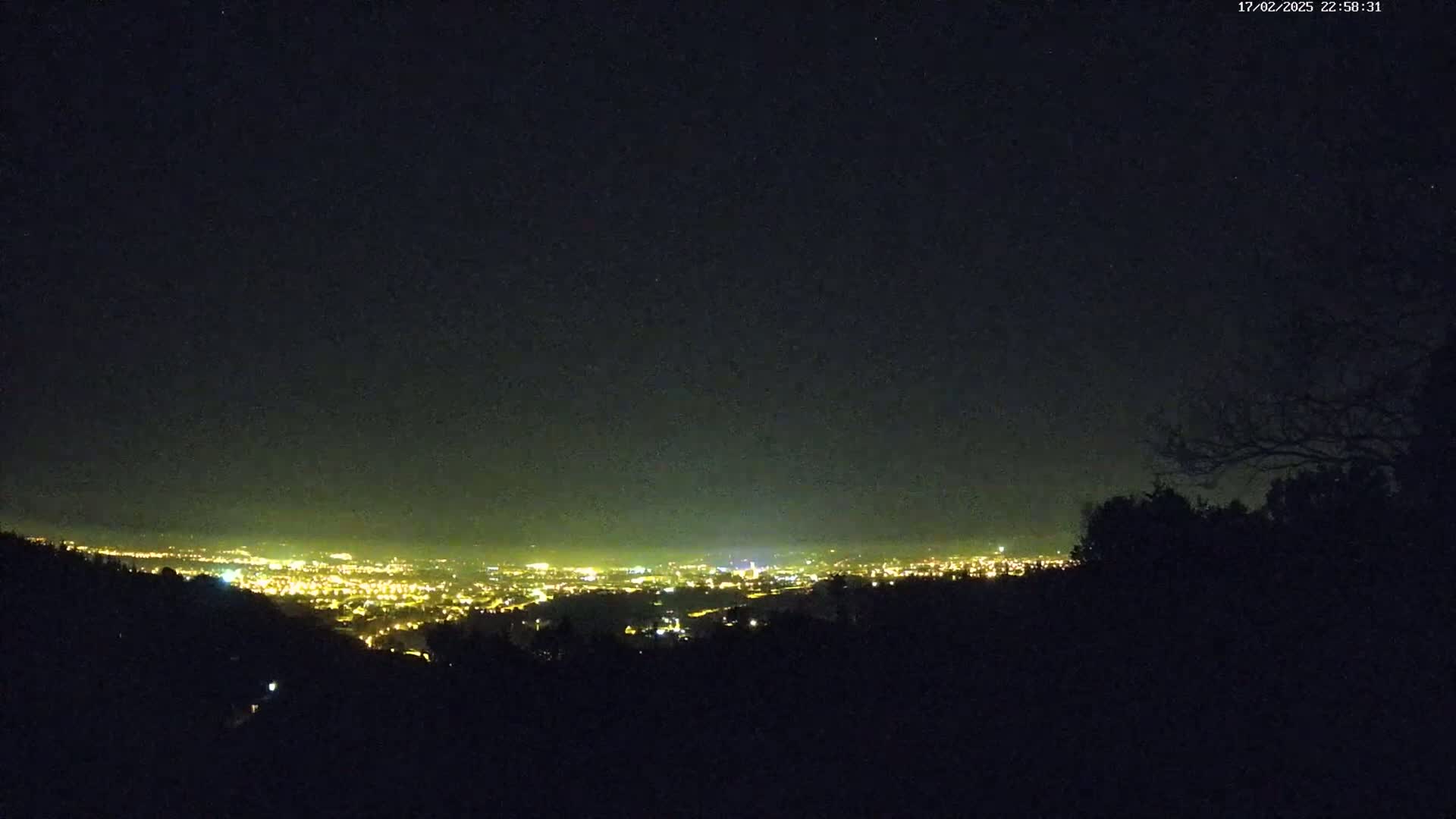 A nighttime view from a hilltop shows a distant city glowing with lights under a clear, dark sky.