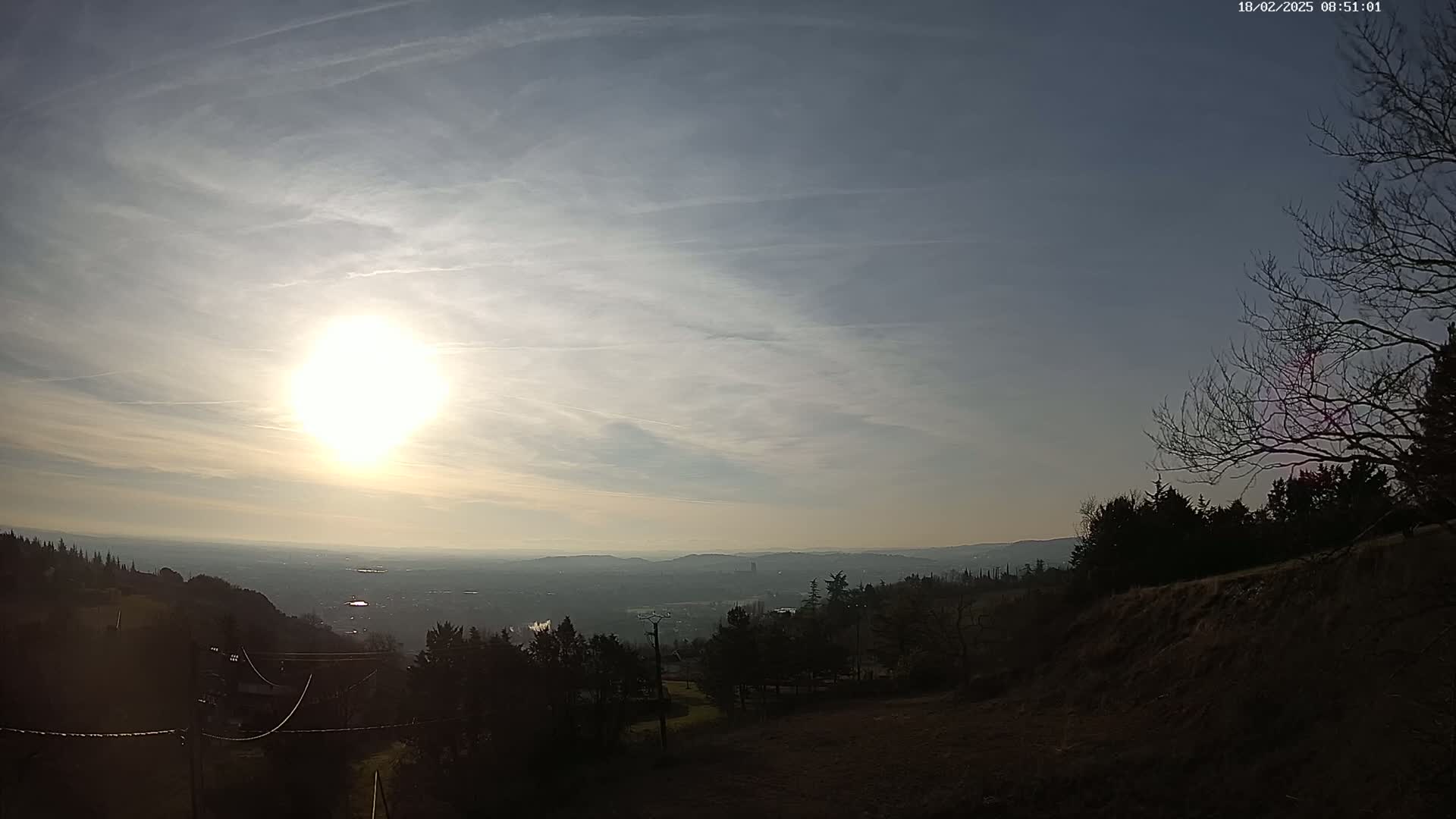 A bright sun shines in a mostly clear sky over a hazy, distant valley and dark, sparsely-treed hillside.