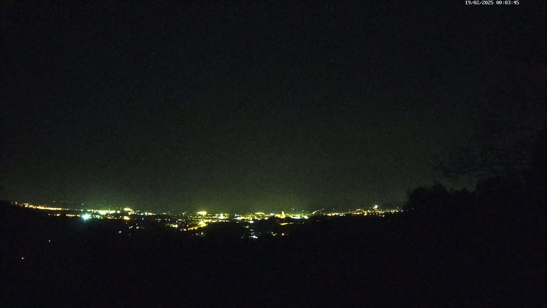 A nighttime view of a distant city's lights spread across a valley, seen from a dark, elevated vantage point under a clear, starry sky.