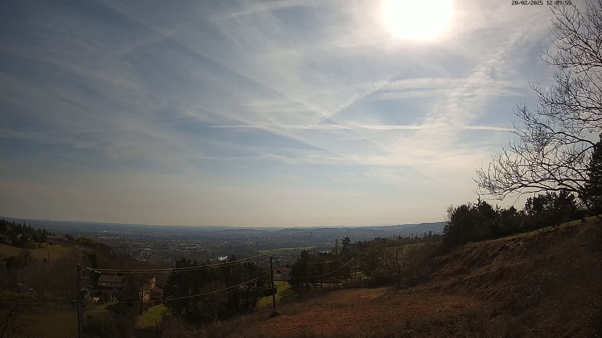 A sunny day with scattered clouds reveals a panoramic view of a valley town from a hillside, where bare trees and shrubs dot the landscape.