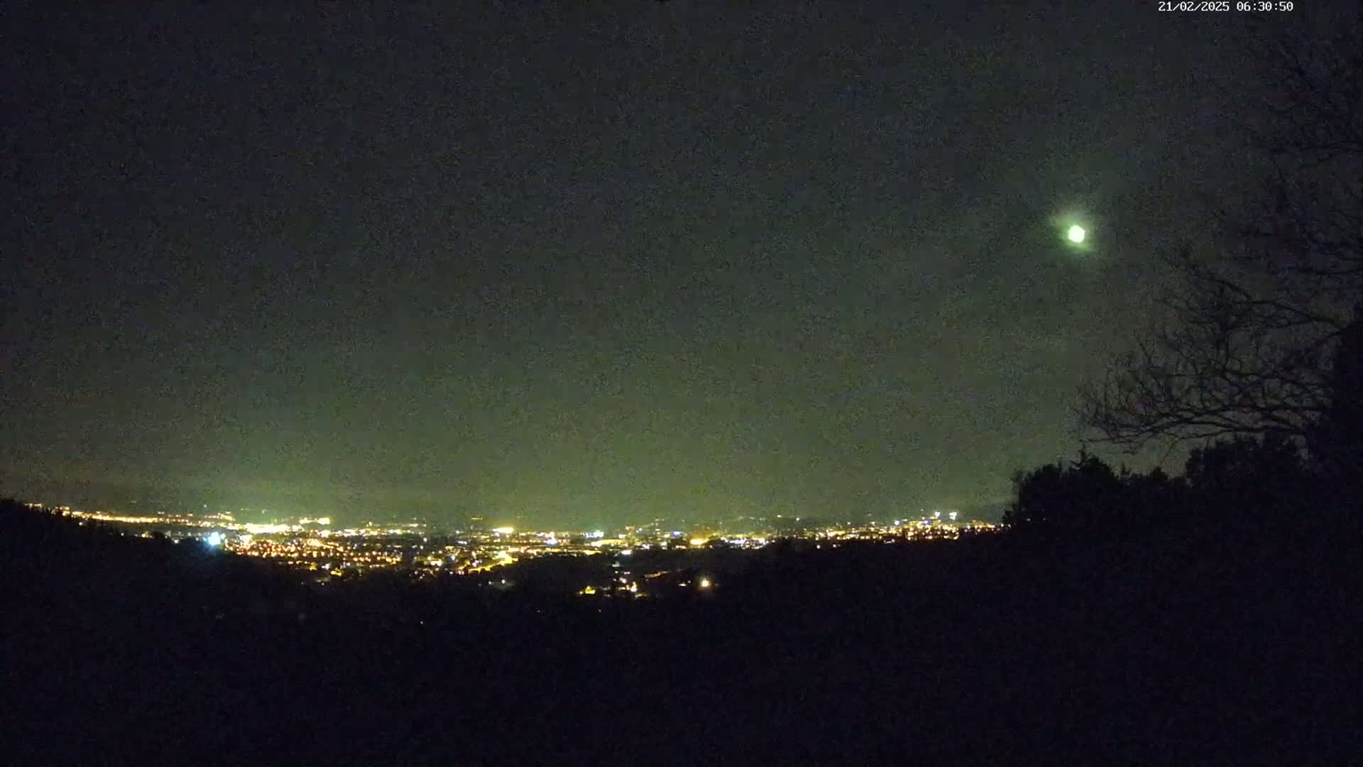 A nighttime view of a city's lights in the distance, partially obscured by a silhouetted tree line, under a dark, possibly cloudy sky with a bright object visible in the upper right.