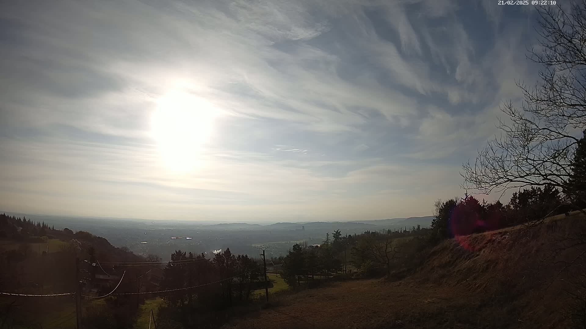 A sunlit, hazy view of a valley and distant city stretches out beneath a partly cloudy sky, from a high vantage point overlooking bare, winter trees and brown earth.
