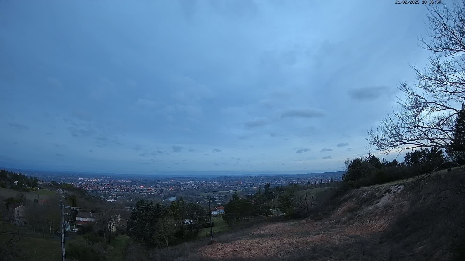 A view from a hilltop at dusk shows a sprawling city lights in the distance under a mostly clear, blue sky.