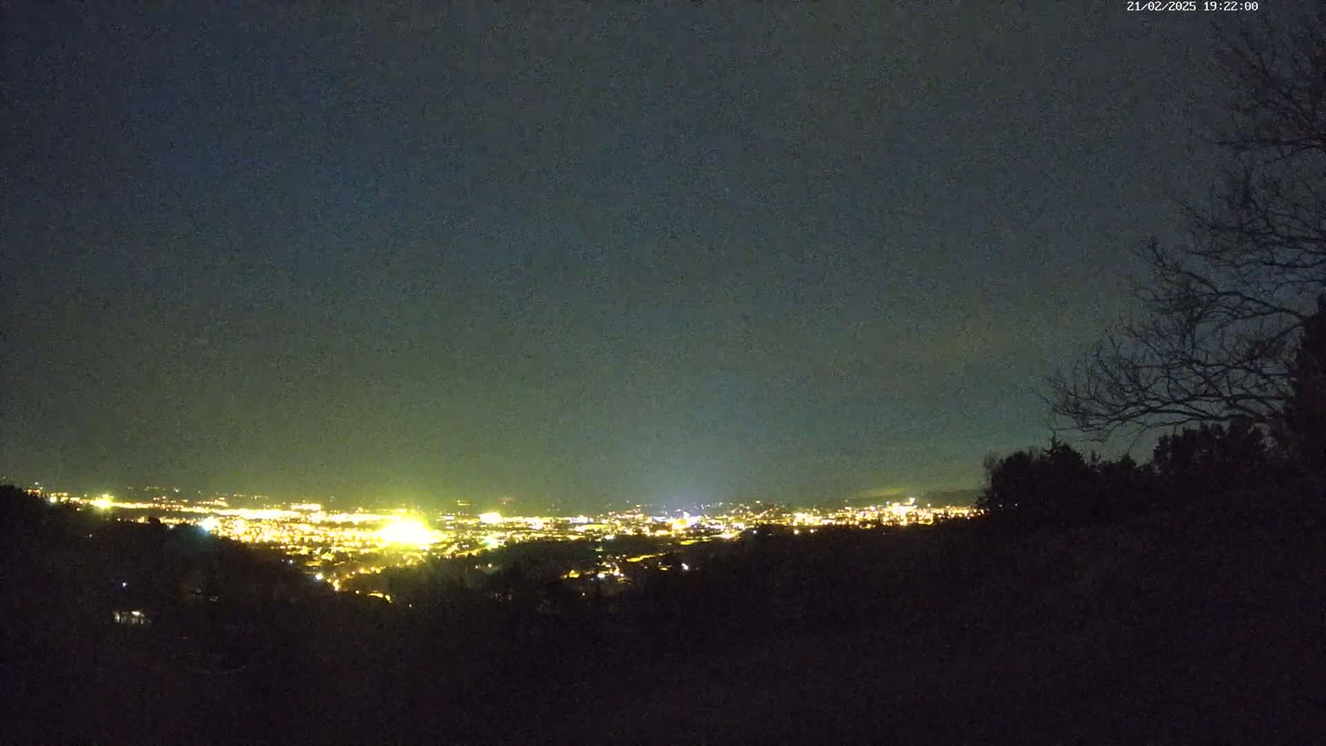 A nighttime view from a hilltop shows a distant city glowing with lights under a dark, clear sky.