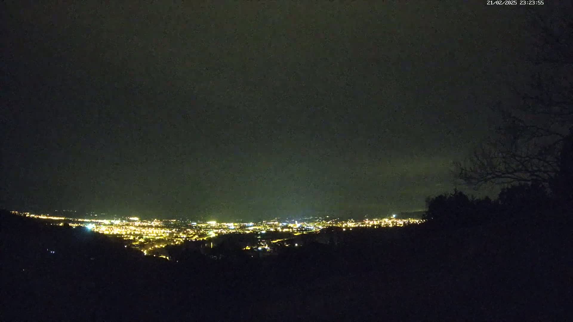A nighttime view of a city's lights spread across a valley, seen from a dark, silhouetted hillside under a cloudy sky.