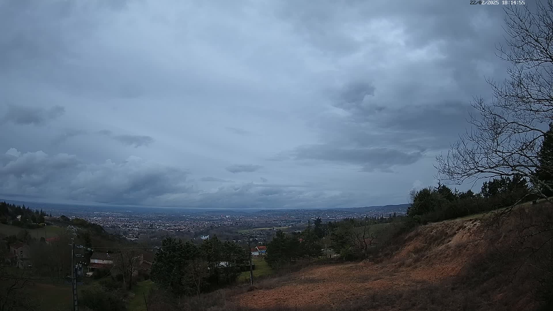 A sprawling city is visible in the distance under a cloudy sky, viewed from a hilly, sparsely vegetated landscape.