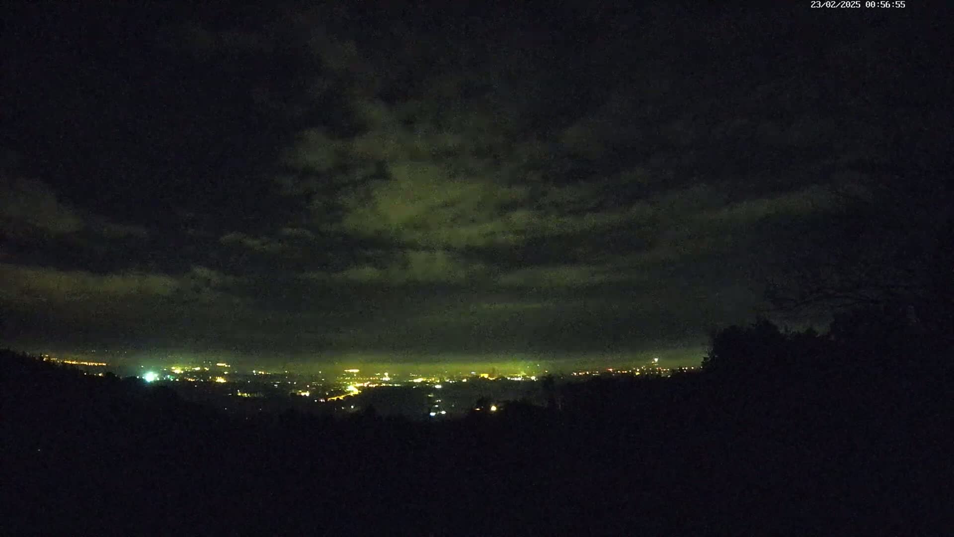 A nighttime view from an elevated position shows a distant city's lights spread across a valley under a cloudy sky.