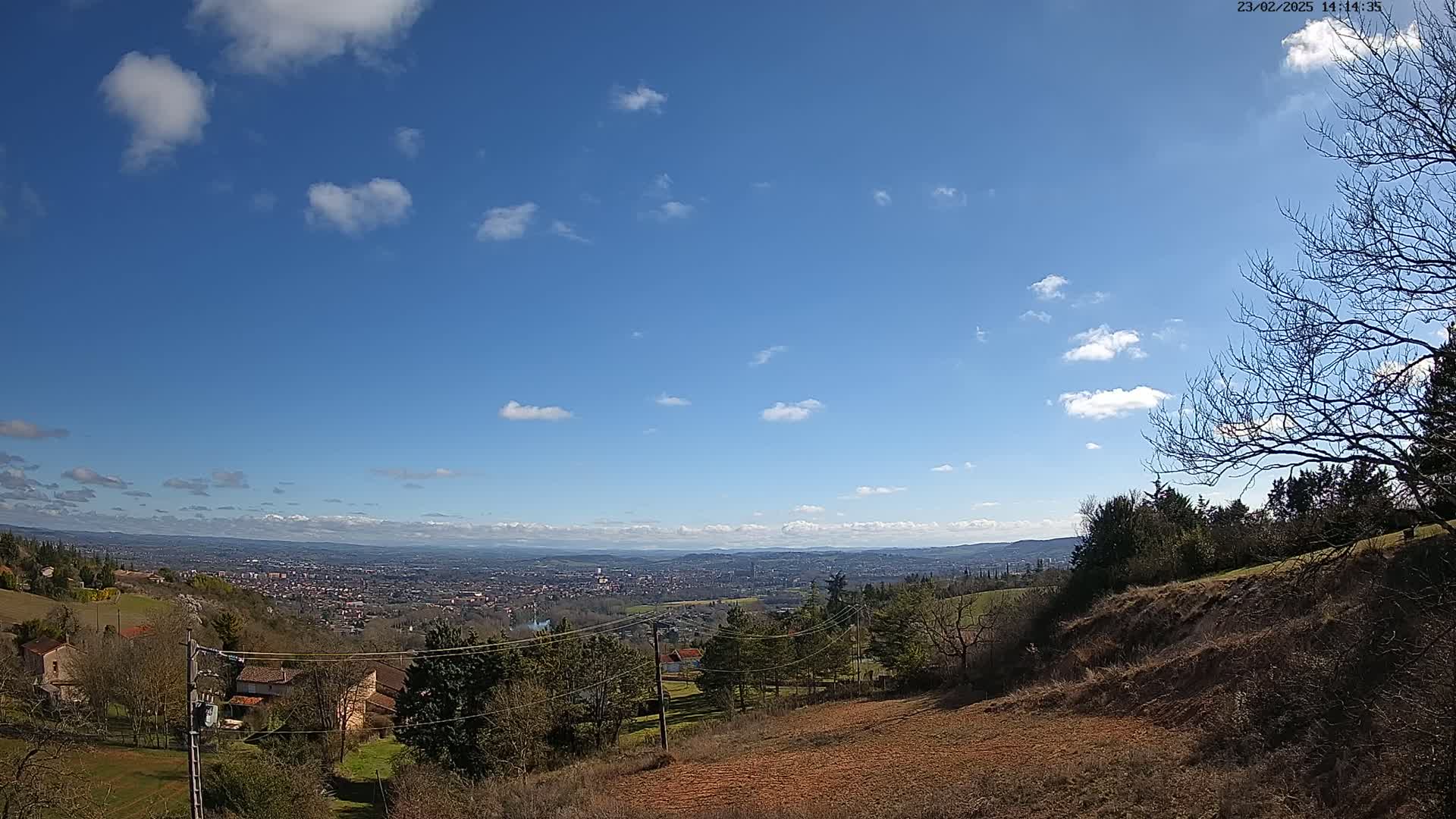 A partly cloudy blue sky overlooks a sprawling landscape of houses, trees, and rolling hills, stretching to a distant city under a bright sun.