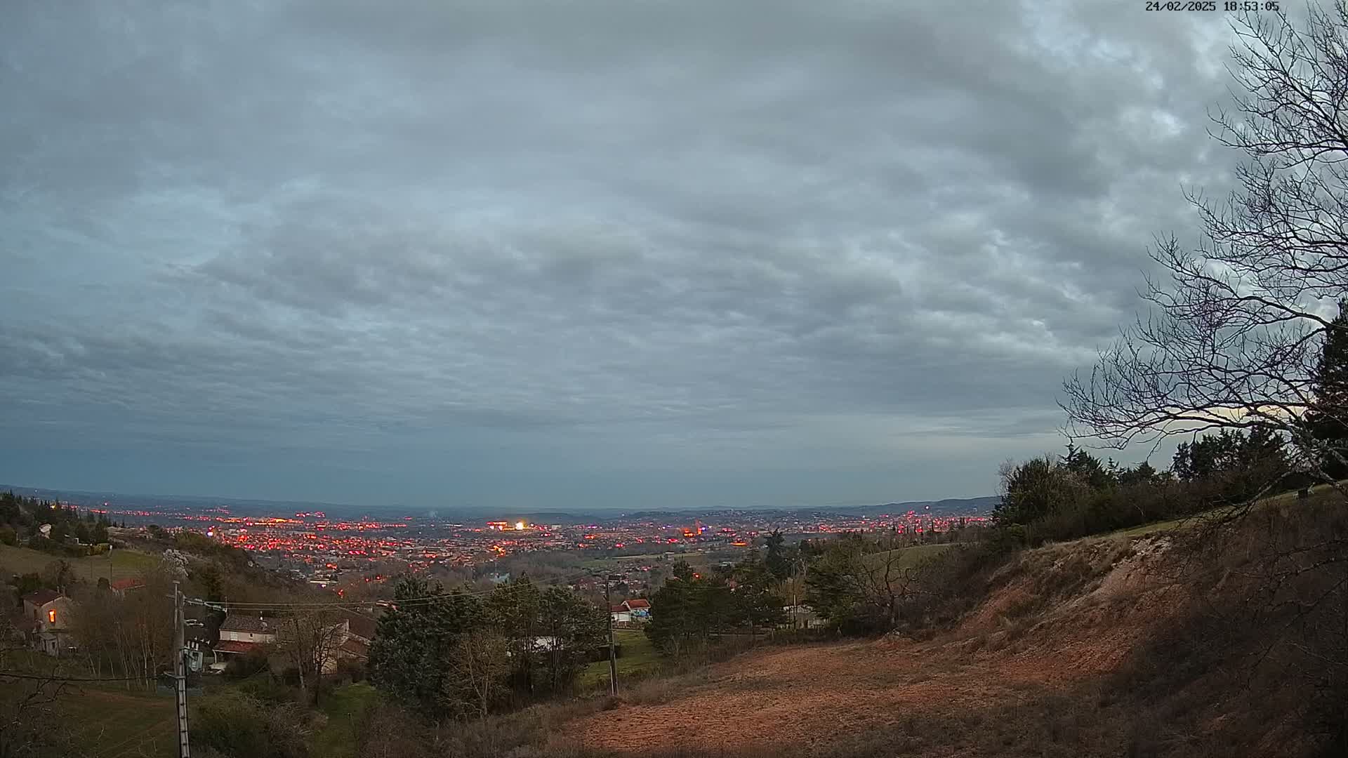 A view of a city at dusk under a cloudy sky, seen from a hilltop with sparse vegetation.