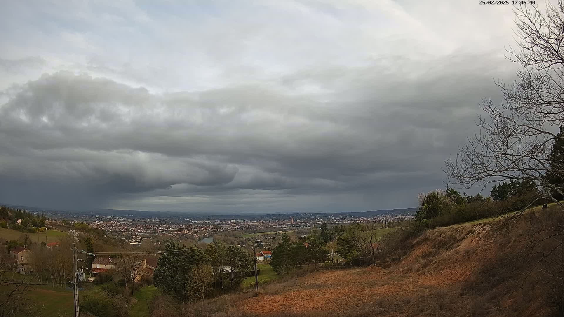 A sprawling town sits in a valley below a hillside covered in sparse, winter trees under a mostly cloudy, gray sky.