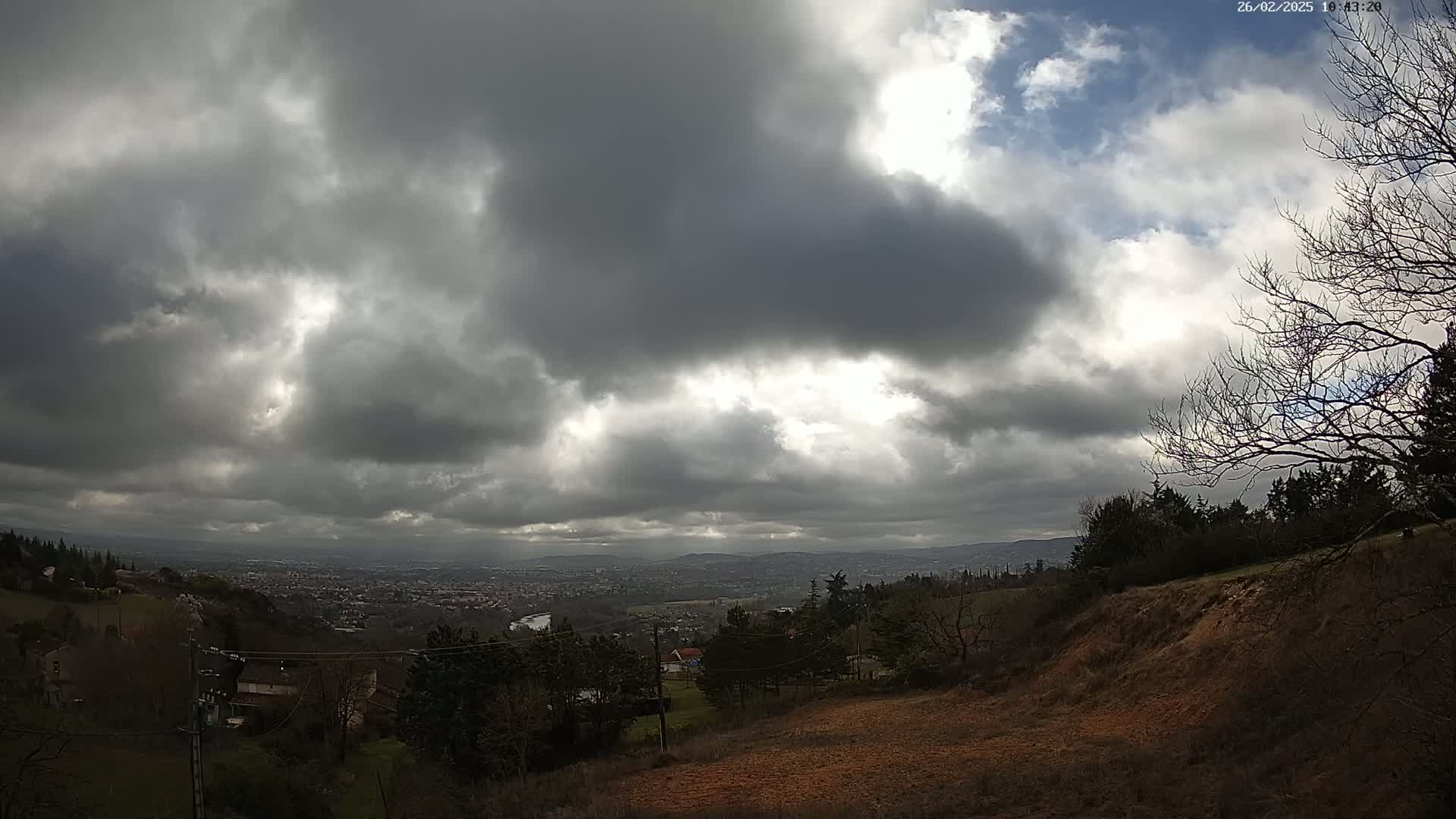 A partly cloudy sky over a hilly landscape featuring a distant city and bare winter trees.