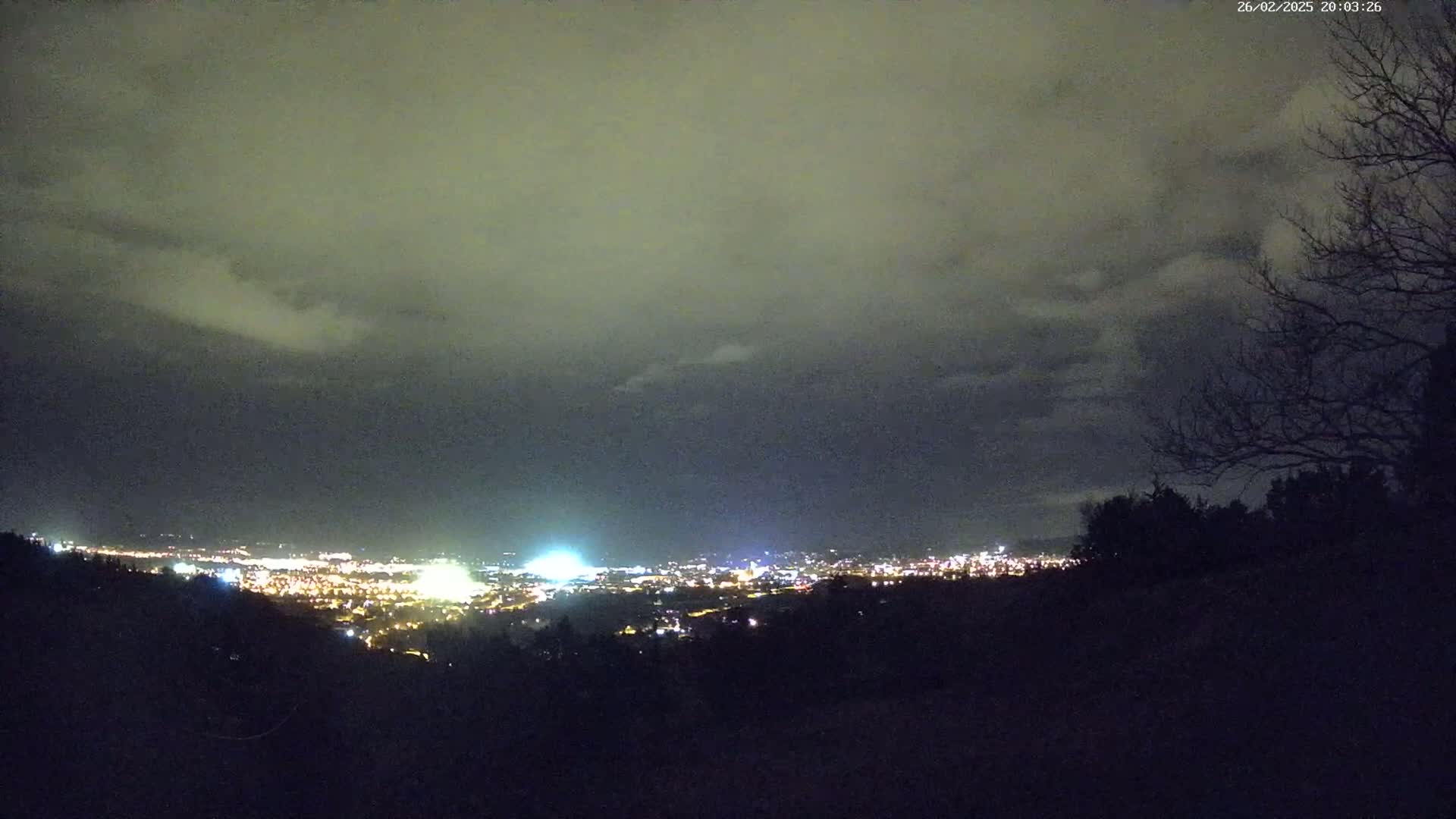 A nighttime view from a hilltop shows a city's lights spread across a valley under a cloudy sky.