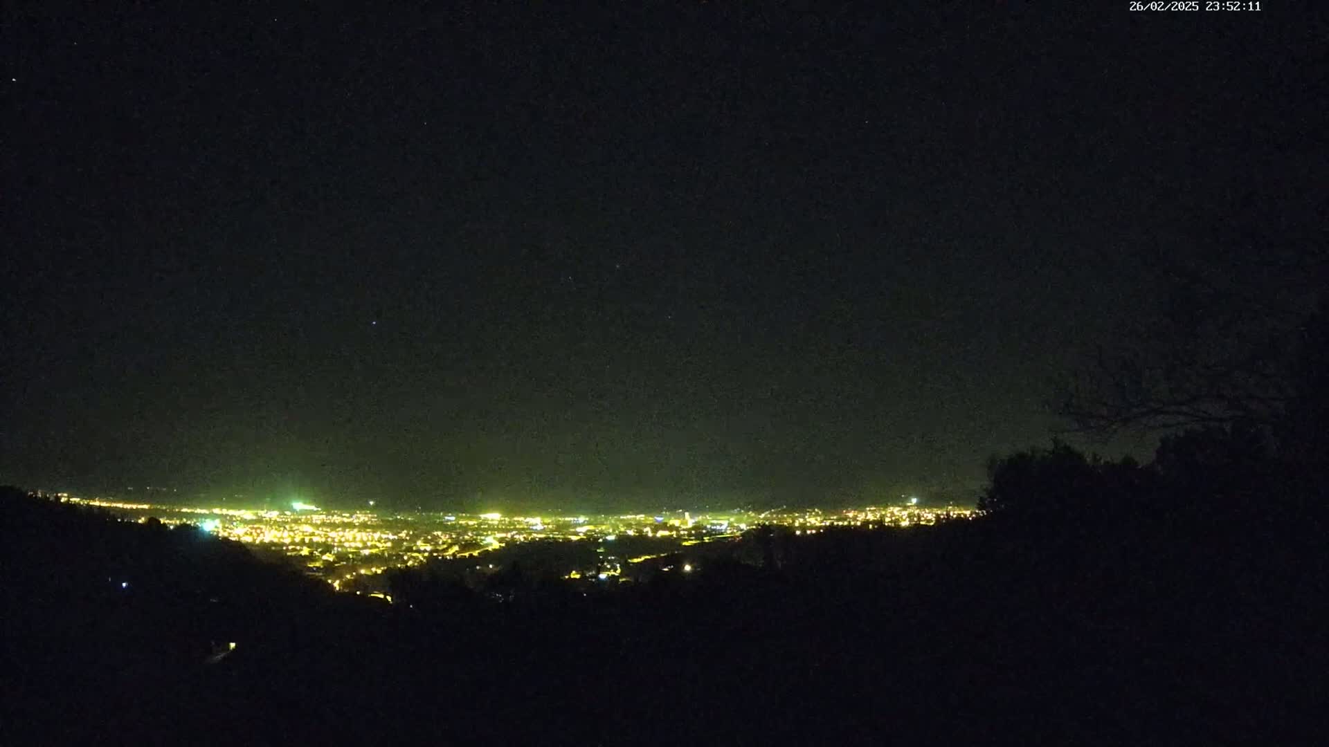 A nighttime view from a hilltop shows a distant city illuminated with many lights under a clear, starry sky.