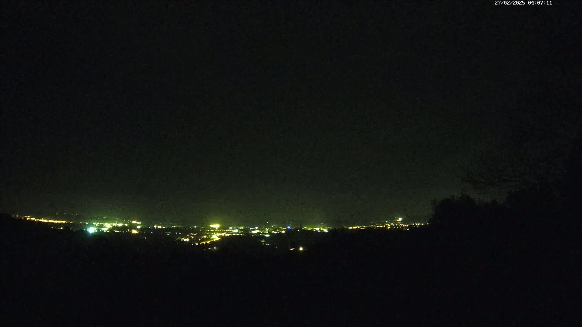 A nighttime view from a hilltop shows a distant city glowing with scattered lights under a dark, clear sky.