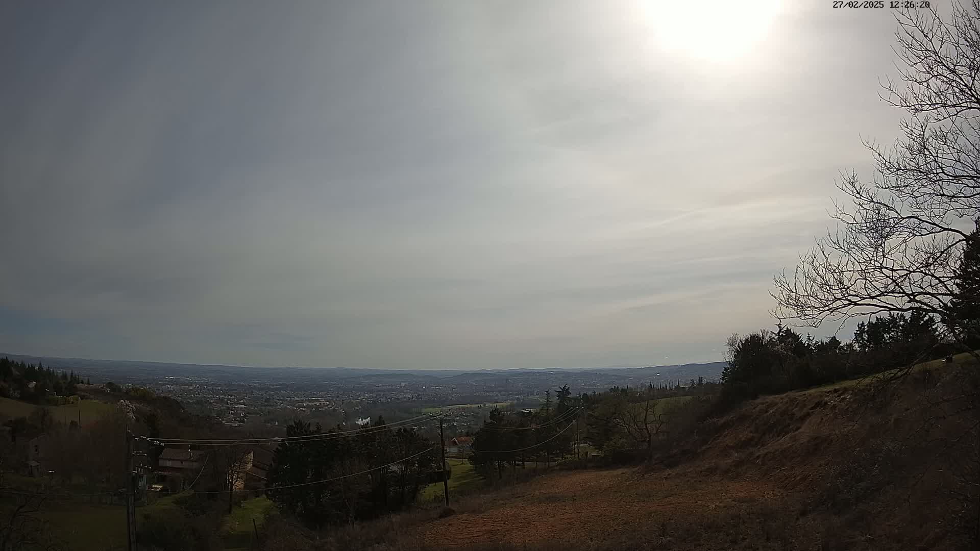 A hazy, sunlit view shows a valley with a distant city below a hillside with bare trees and scrub brush.