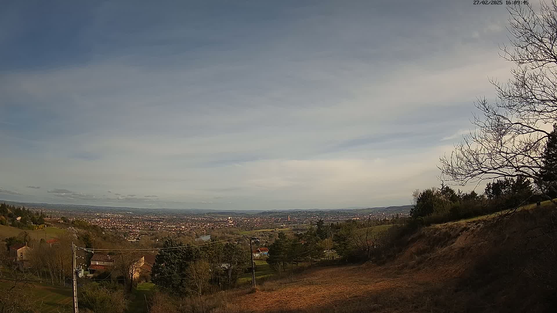 A sprawling city view is seen from a hillside under a mostly clear, light blue sky.