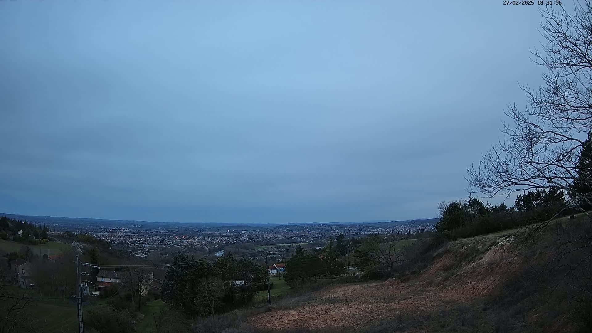 A panoramic view of a sprawling city at dusk, seen from a hilly, sparsely wooded area under a mostly clear, pale blue sky.