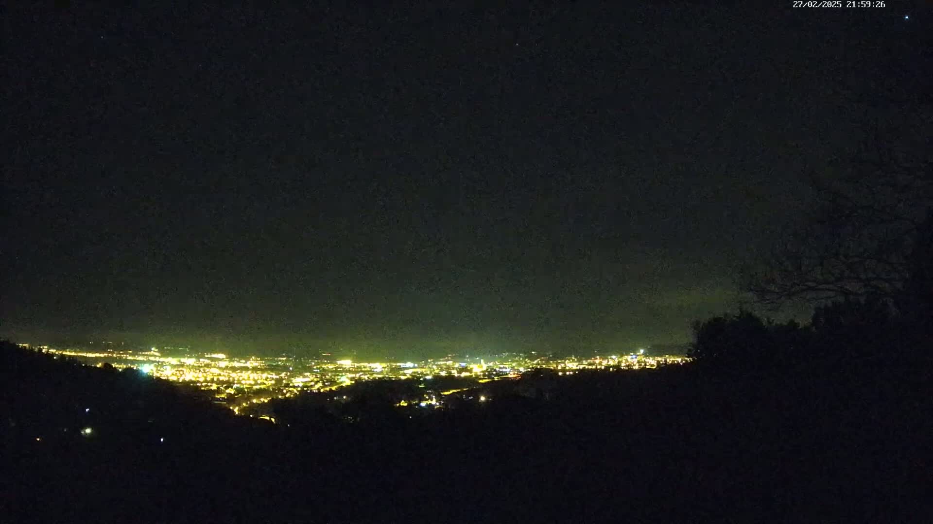 A nighttime view from a hilltop shows a city's lights spread across a valley under a clear, dark sky.