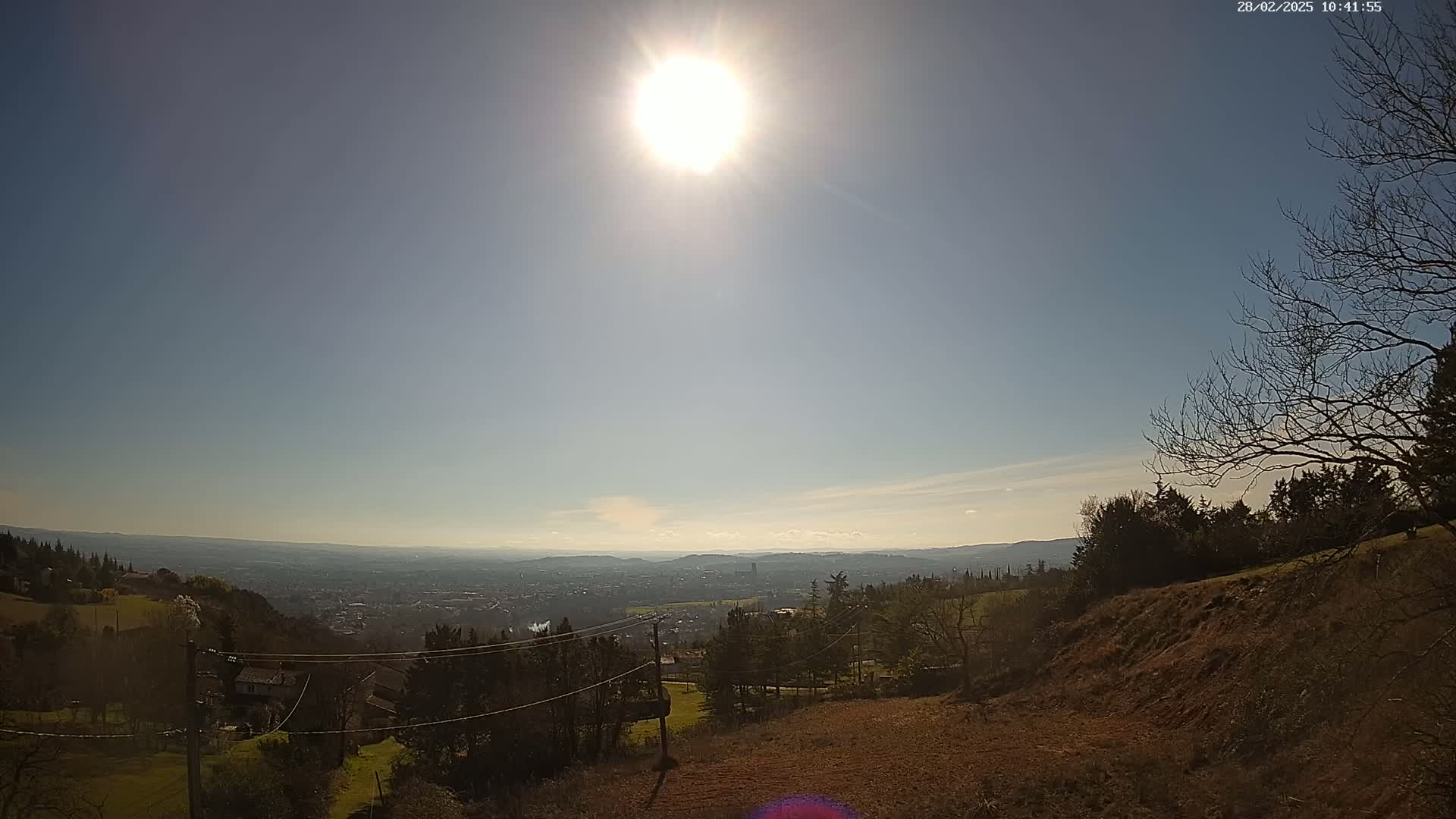 A sunny, clear day reveals a sprawling vista of a city nestled in a valley, viewed from a high vantage point with sparse trees and brown earth in the foreground.