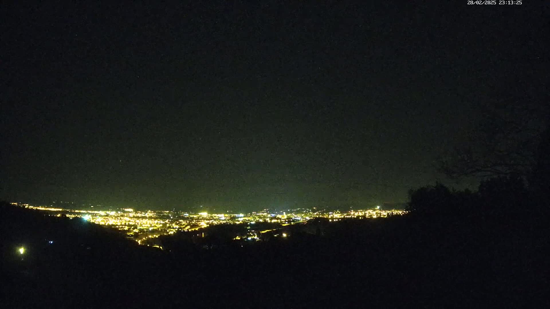A nighttime view from a hilltop shows a distant city glowing with lights under a clear, starry sky.