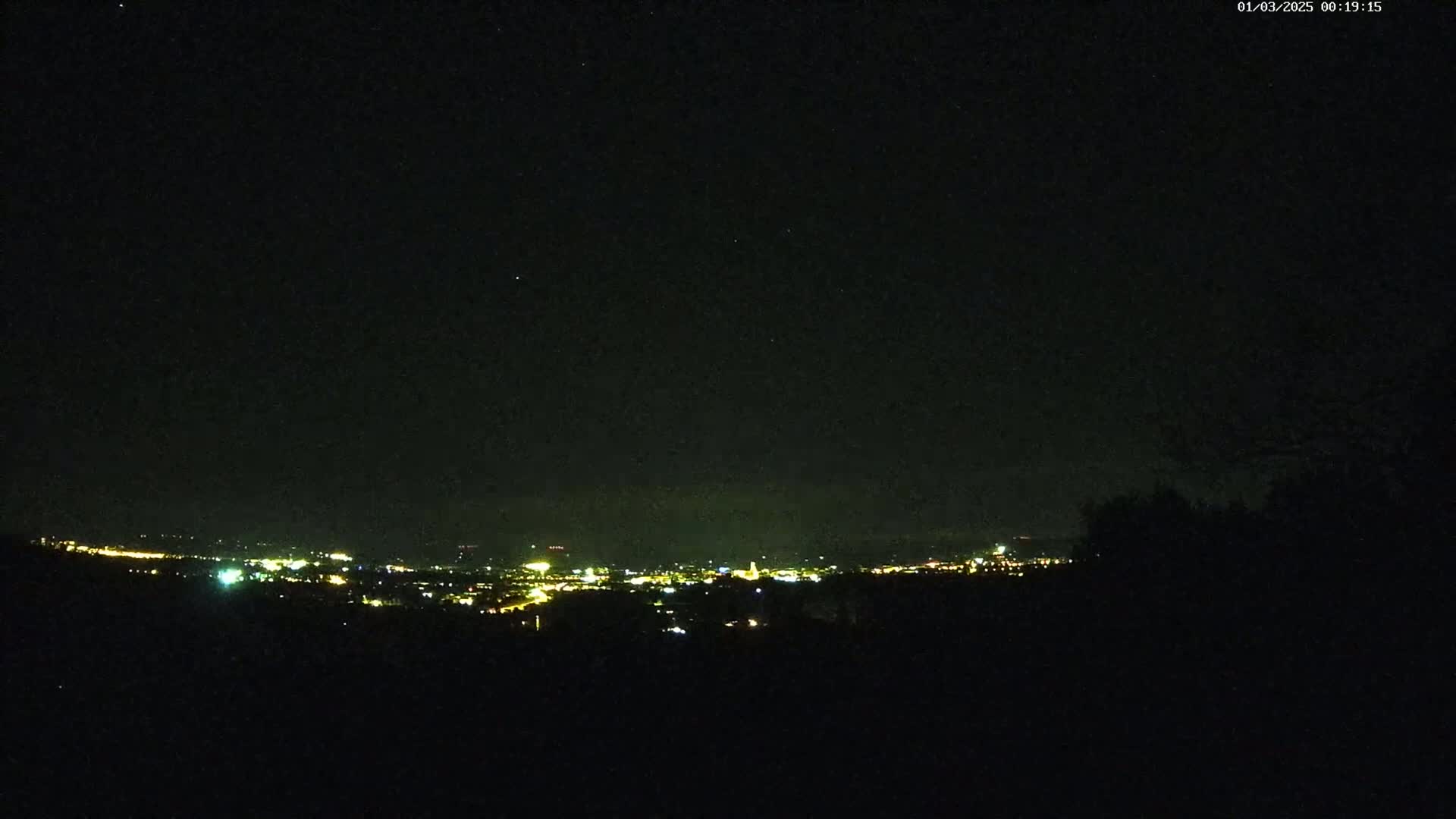 A nighttime, clear sky view of a distant city illuminated by lights, seen from a dark, elevated vantage point.