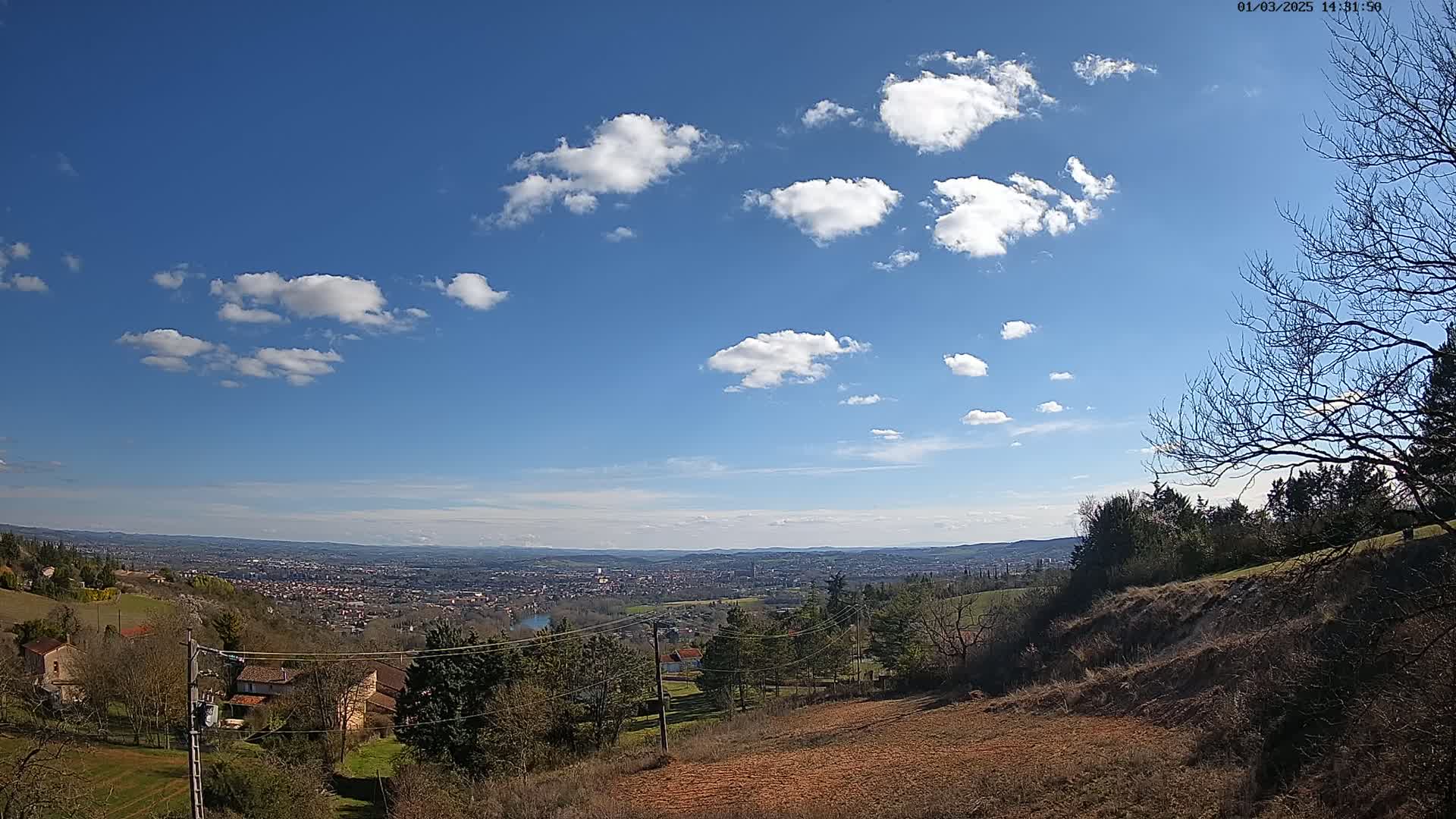 A sunny day with scattered cumulus clouds reveals a panoramic view of a sprawling city nestled in a valley, surrounded by rolling hills and sparsely wooded areas.