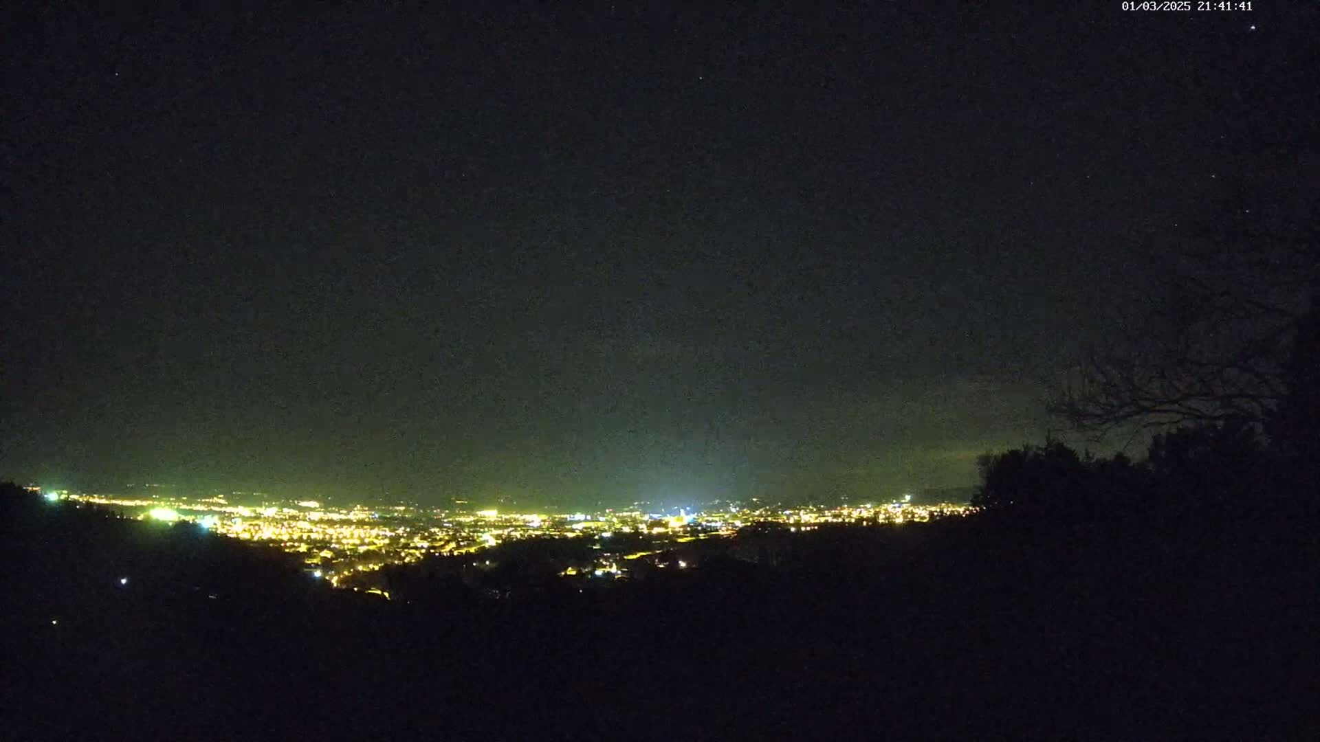 A nighttime view from a hilltop shows a distant city glowing with lights under a clear, dark sky.