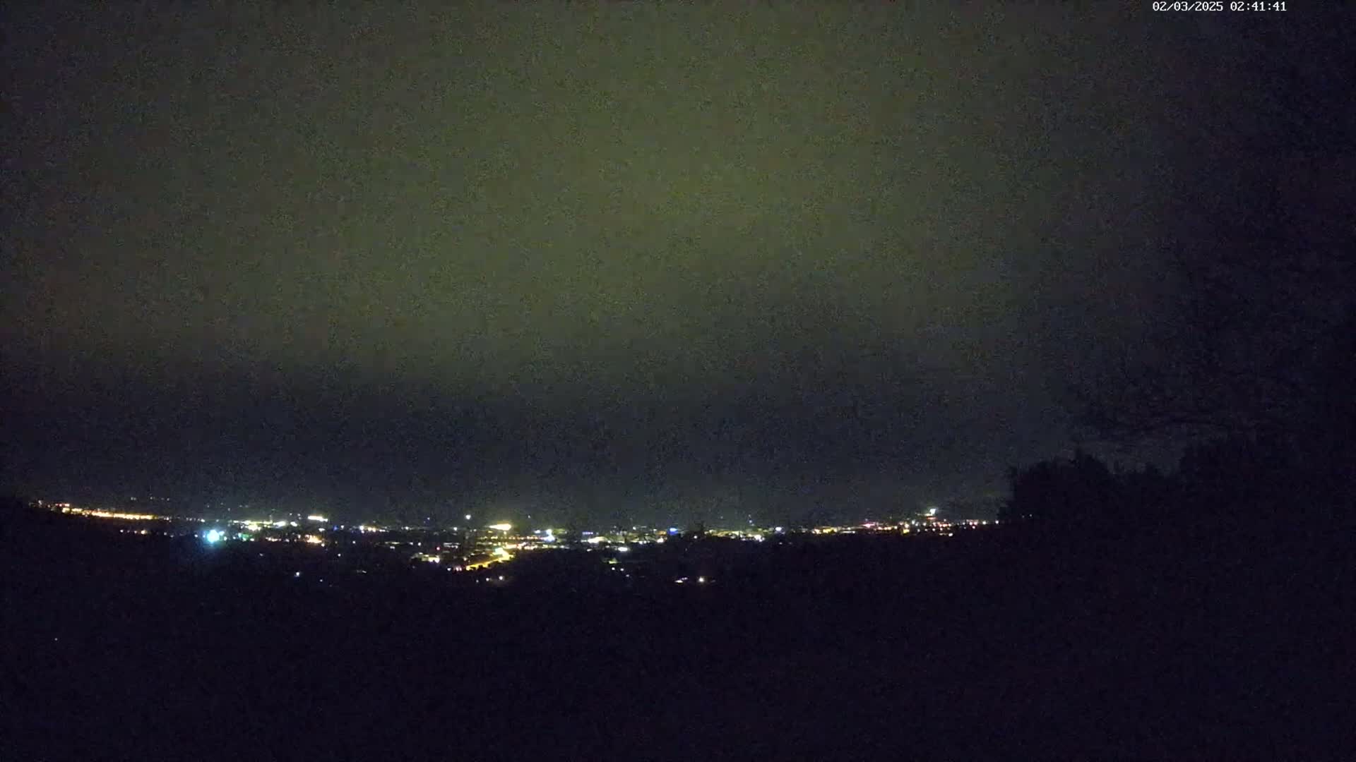 A nighttime view of a city skyline illuminated by distant lights under a dark, overcast sky.