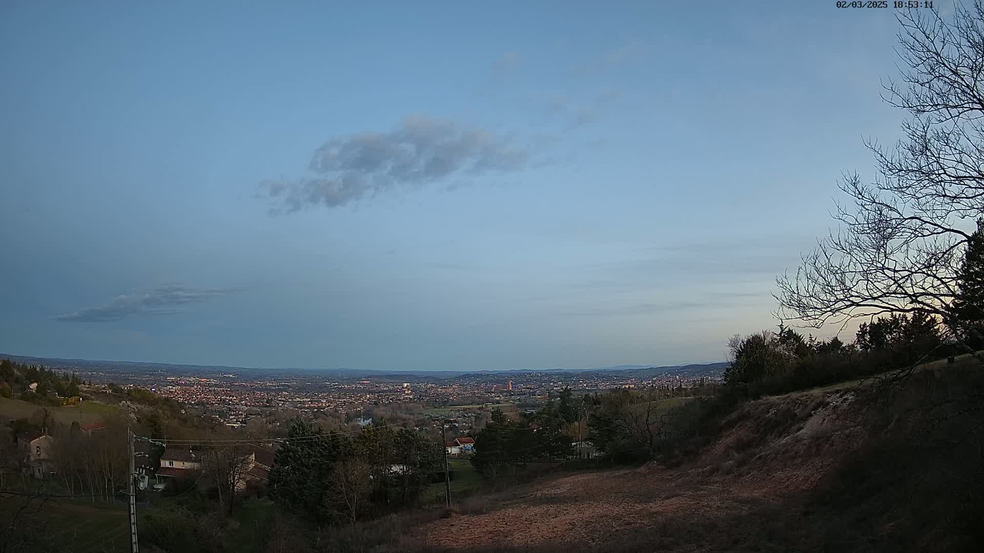 A sprawling city is visible in the distance under a mostly clear, twilight sky, viewed from a hillside dotted with leafless trees and houses.