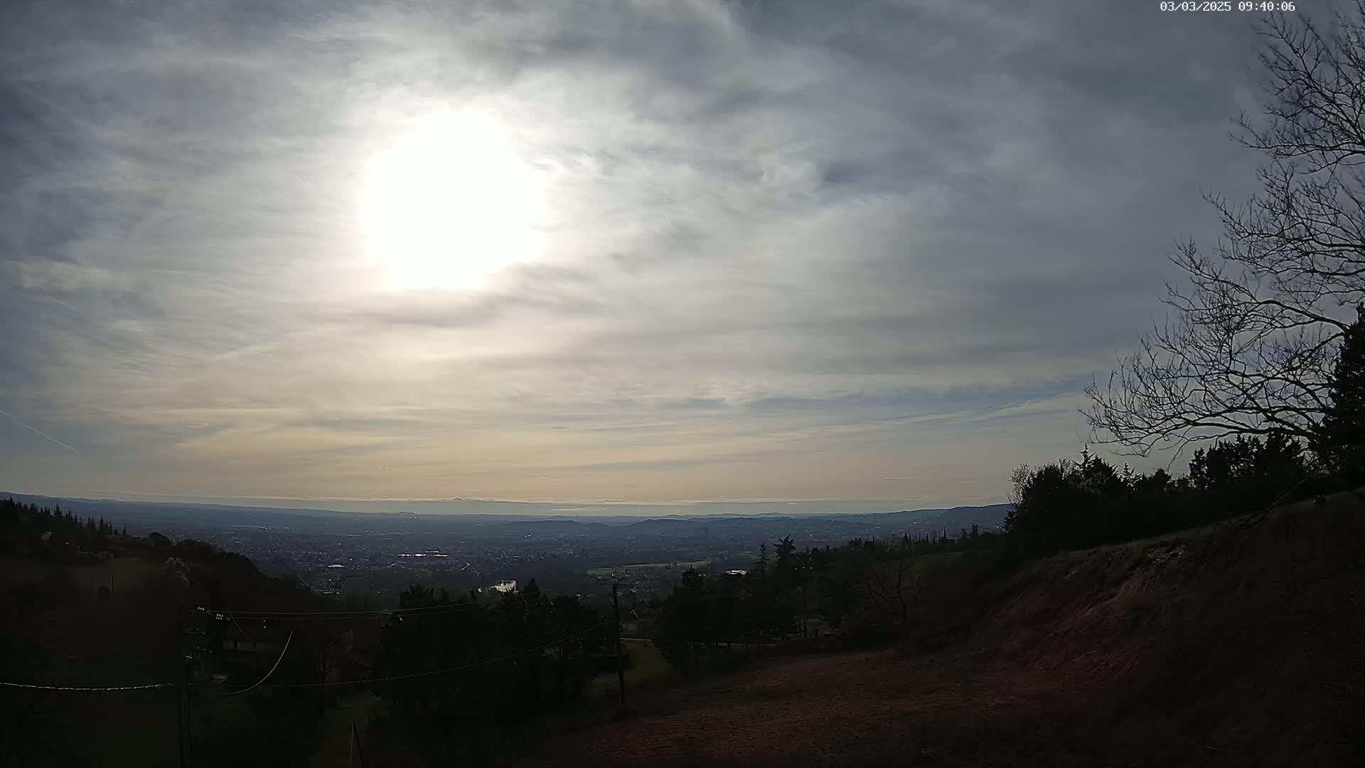 A view from a hilltop shows a partly sunny sky over a distant city and valley, with bare trees in the foreground.