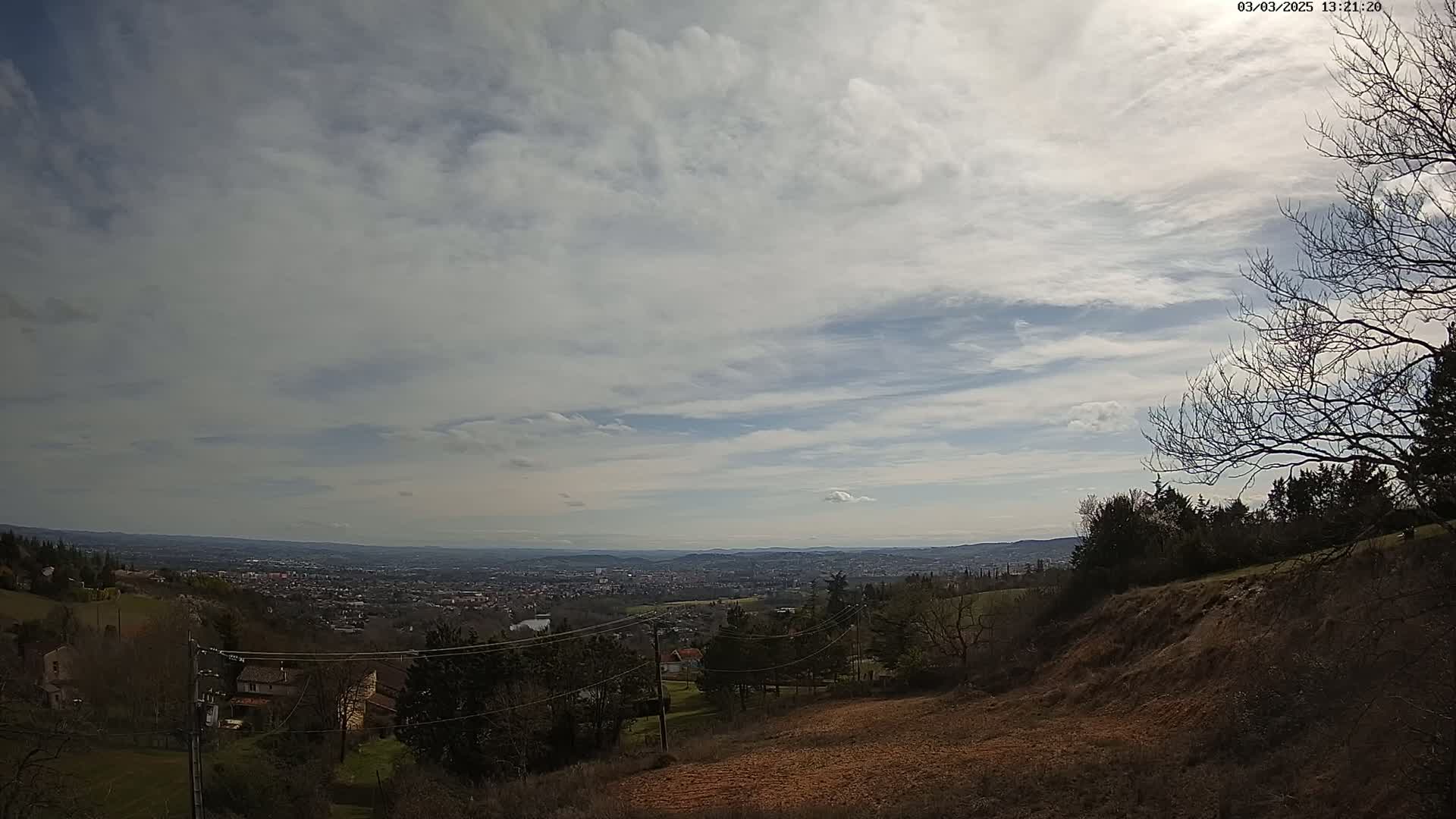 A partly cloudy sky overlooks a sprawling city and hilly landscape, with leafless trees and a barren hillside in the foreground.