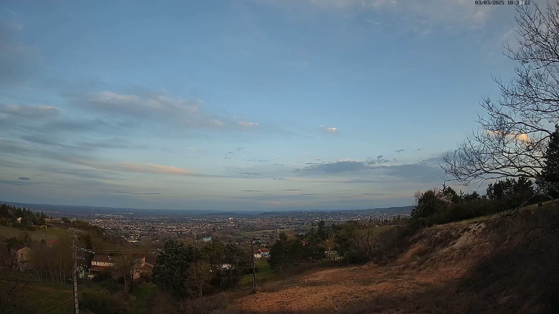 A hillside overlooking a sprawling city under a mostly clear, light-blue sky with scattered clouds at dusk.