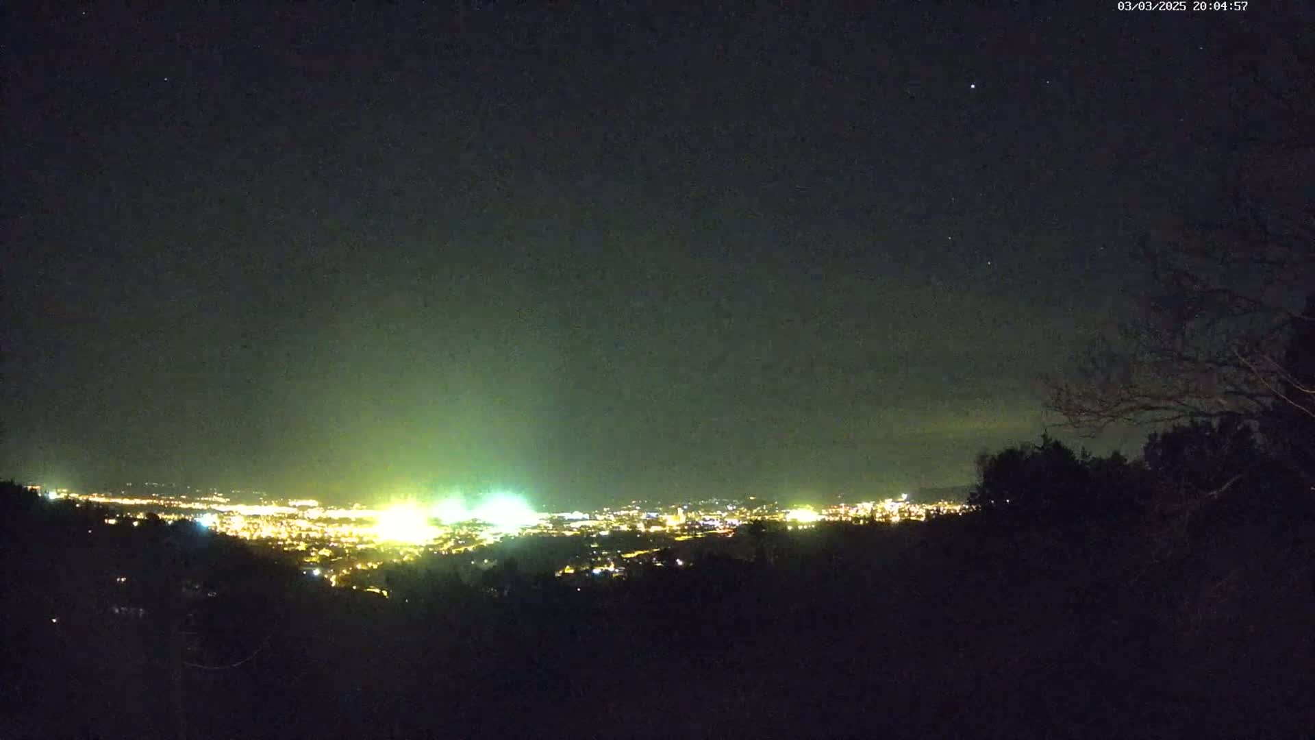 A nighttime view from an elevated position shows a city's lights spread across a valley under a clear, starry sky.