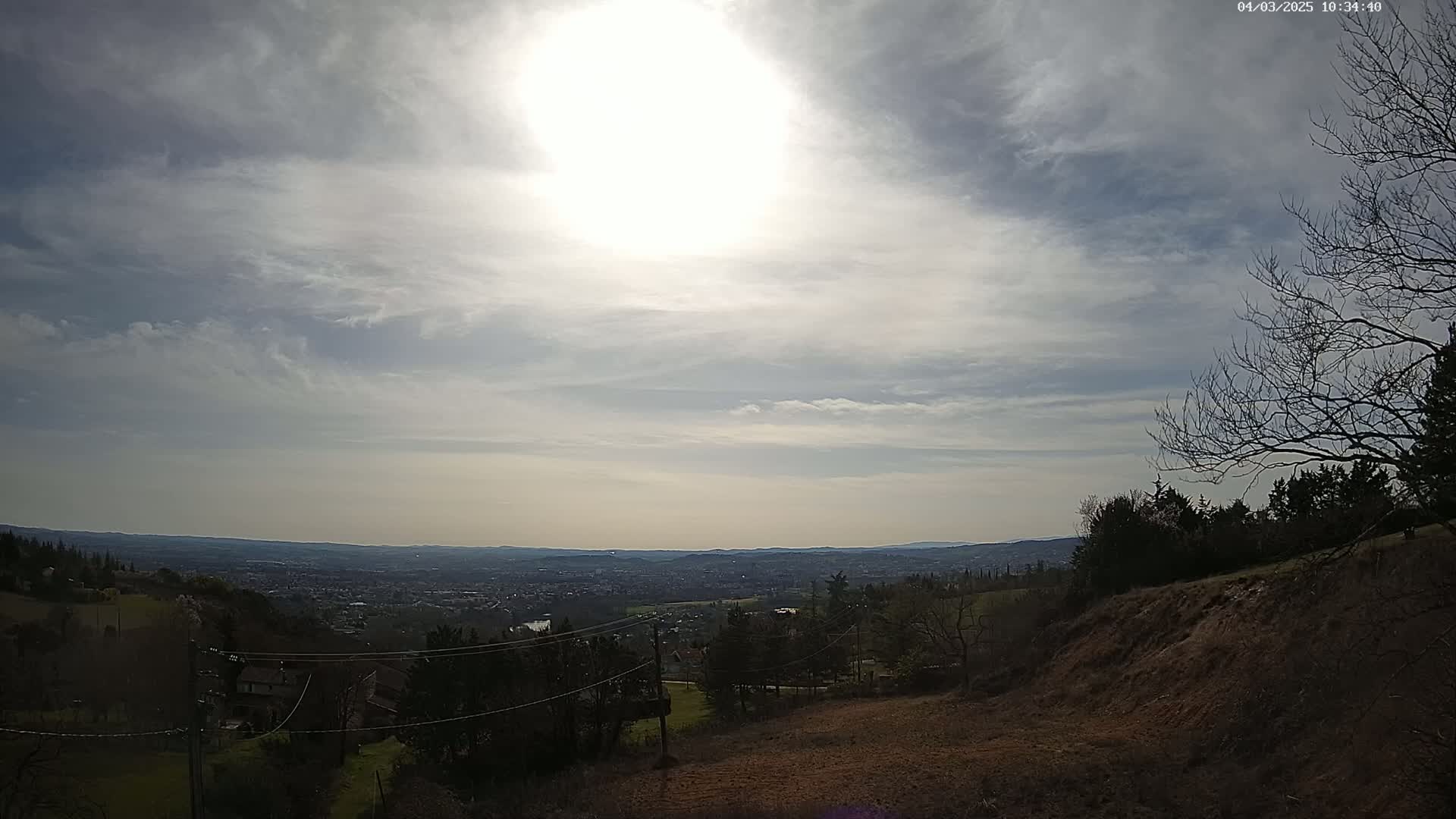 A partly cloudy sky brightens a view of a distant city nestled in a valley, seen from a hillside with bare trees and sparse vegetation.