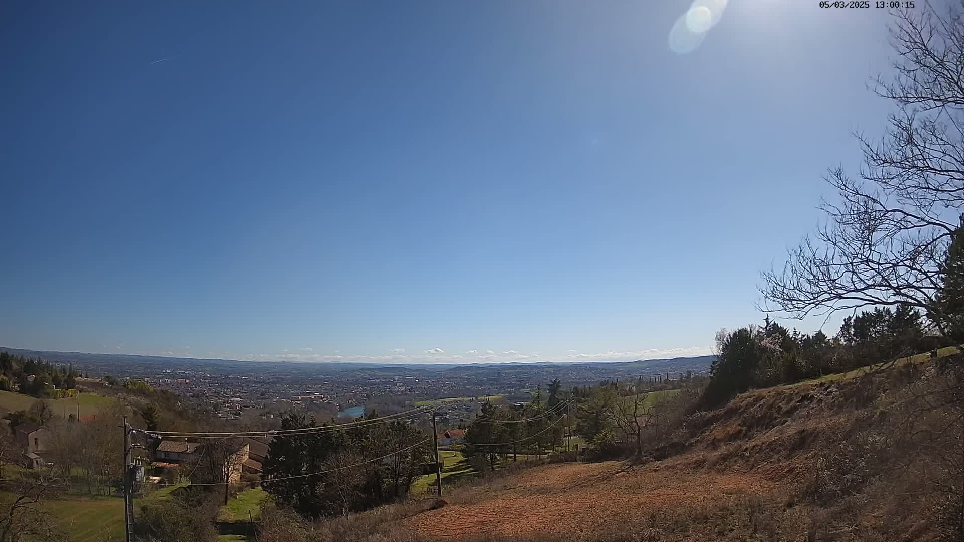 A sunny day reveals a panoramic view of a distant city nestled in a valley, seen from a hillside with sparse trees and brown earth in the foreground.