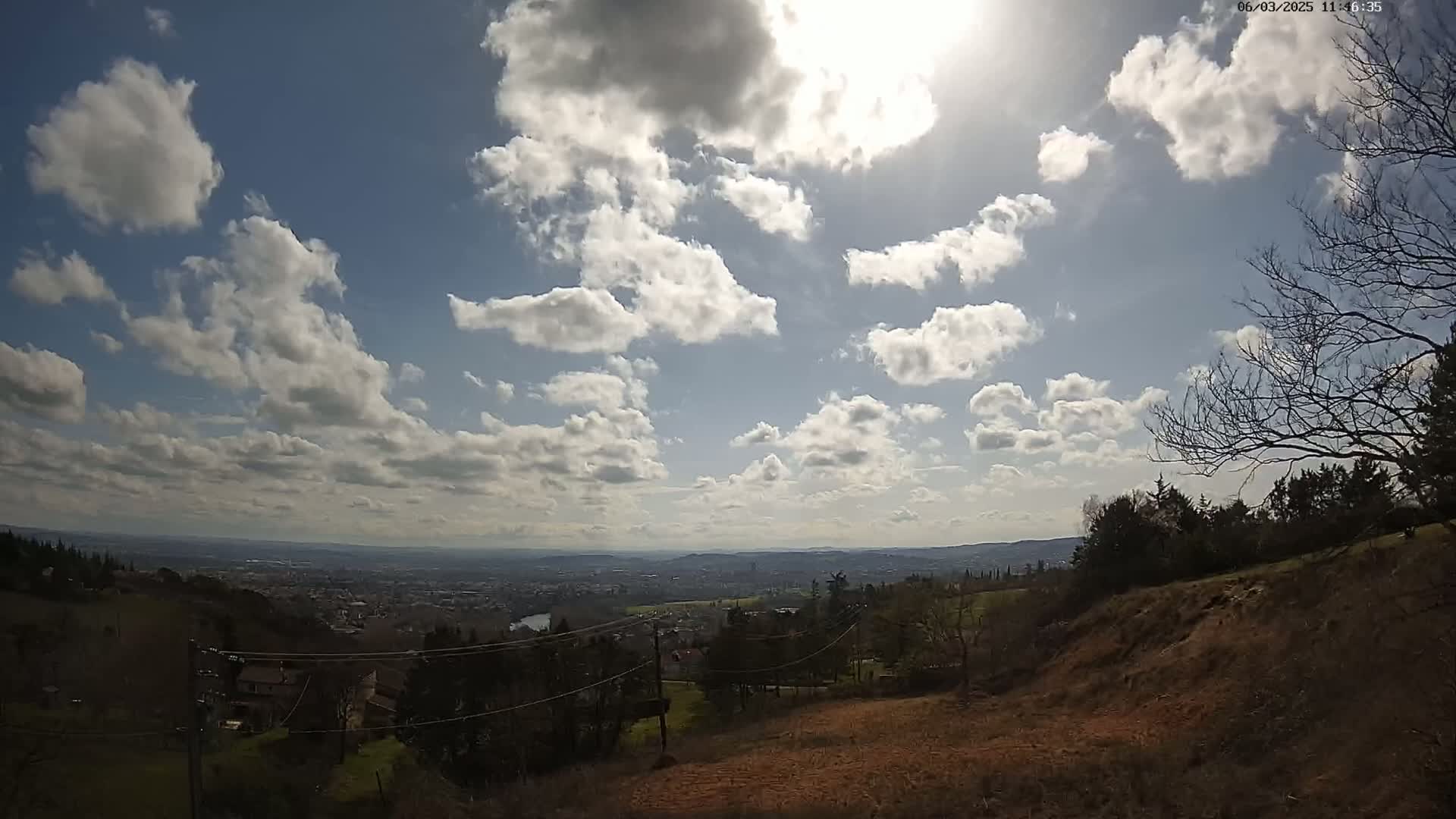 A sunny day with scattered cumulus clouds overlooks a sprawling valley and distant city, viewed from a hillside with bare trees and brown earth.