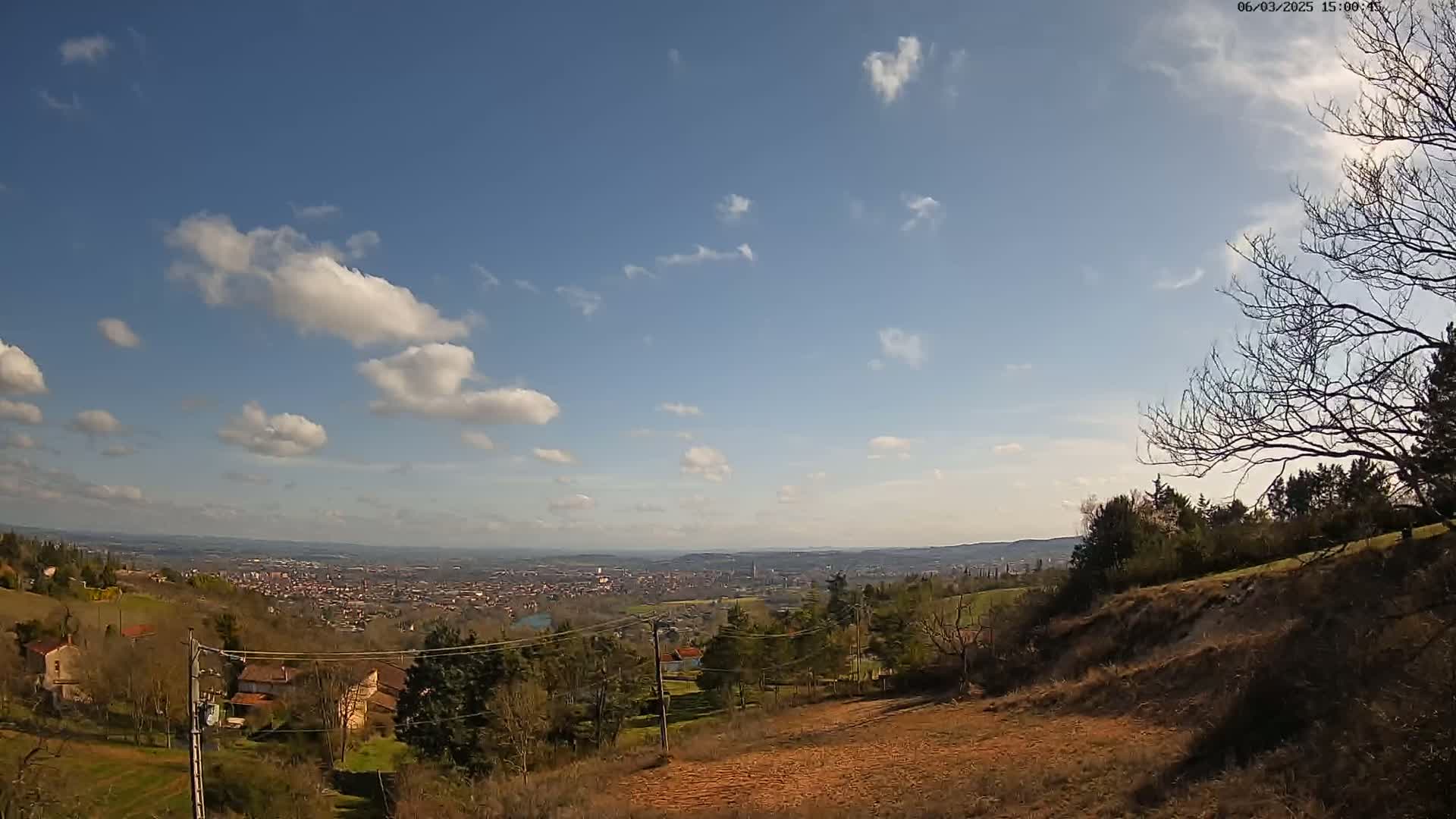 A sprawling view of a valley town stretches beneath a mostly clear blue sky dotted with fluffy white clouds, from a hilly vantage point with bare trees and sparsely populated houses.