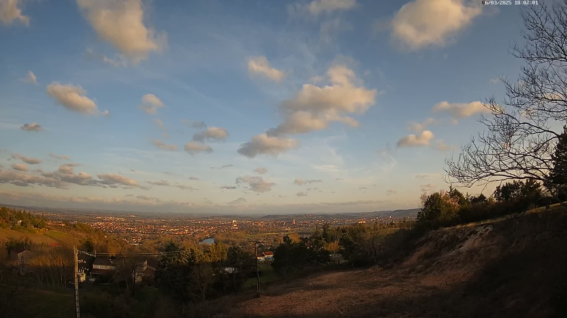 A partly cloudy sunset over a sprawling town nestled in a valley, viewed from a hillside with bare trees and scrubby vegetation.