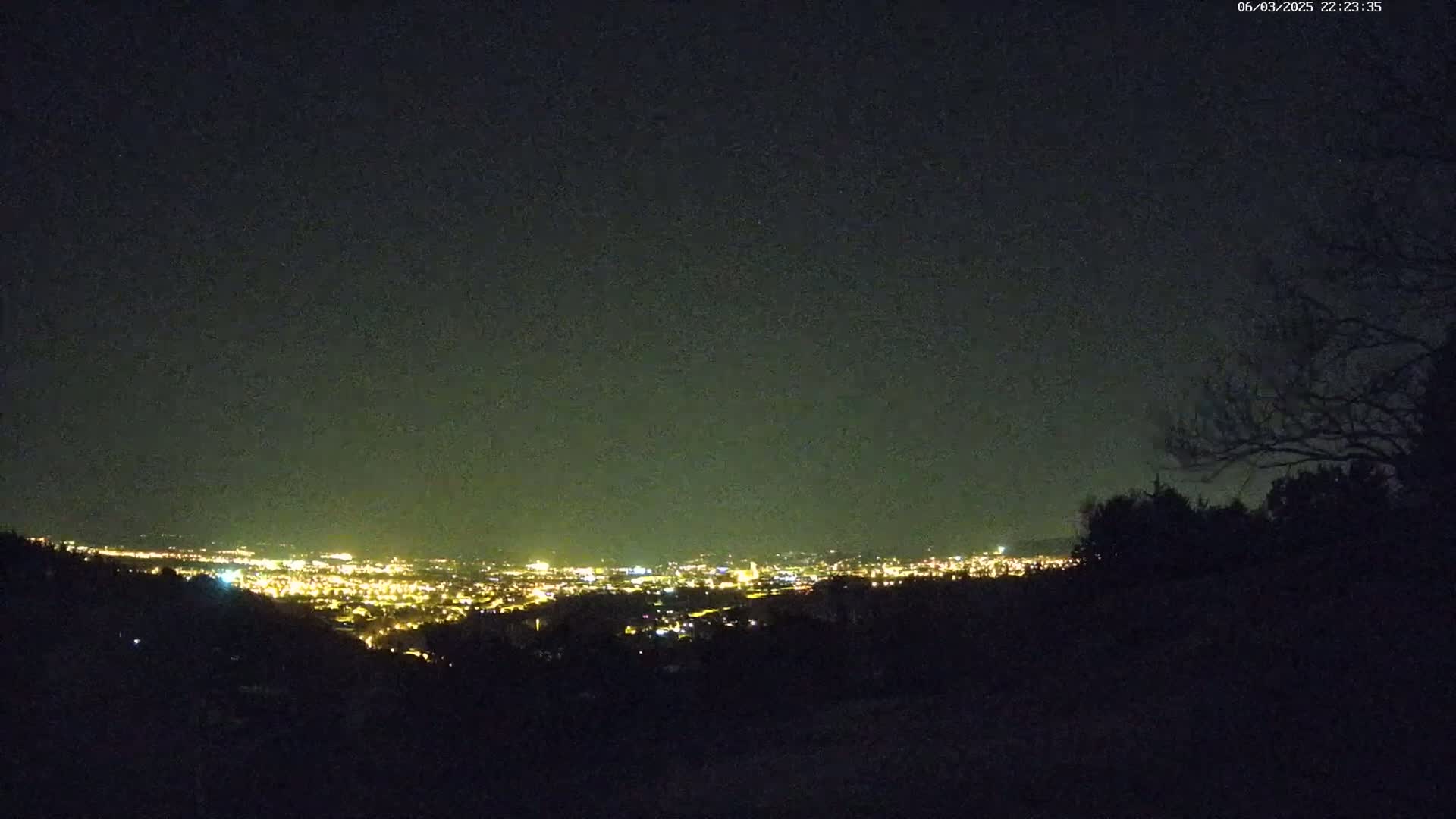 A nighttime view from a hilltop shows a distant city glowing with lights under a dark, clear sky.