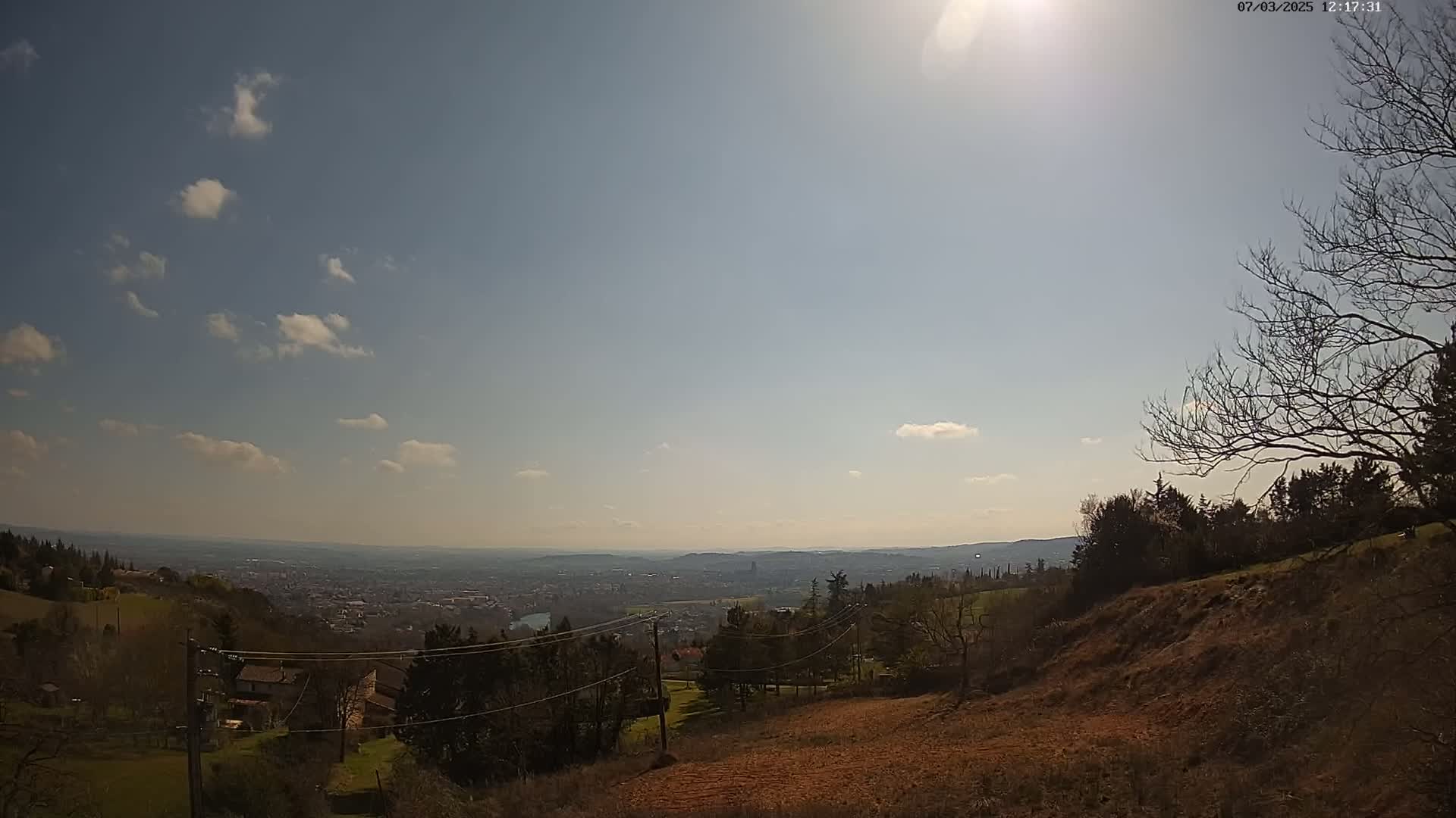 A sunny day with a few clouds shows a panoramic view of a city in the distance from a hillside with bare trees and some buildings.