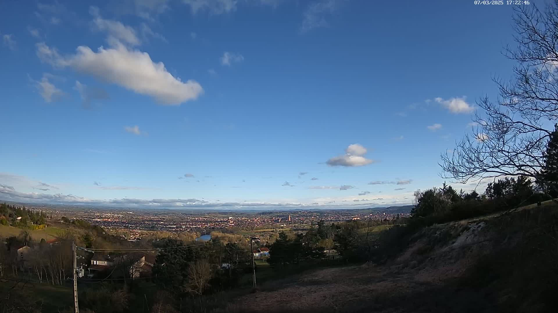 A sprawling city view stretches out under a mostly clear, blue sky dotted with fluffy white clouds, visible from a hillside with sparse trees and buildings.