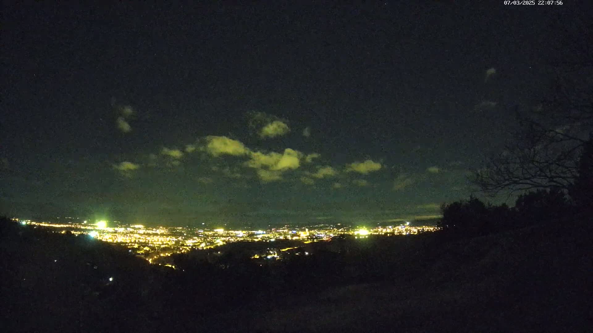 A nighttime view from a hilltop shows a city's lights spread across a valley under a partly cloudy sky.
