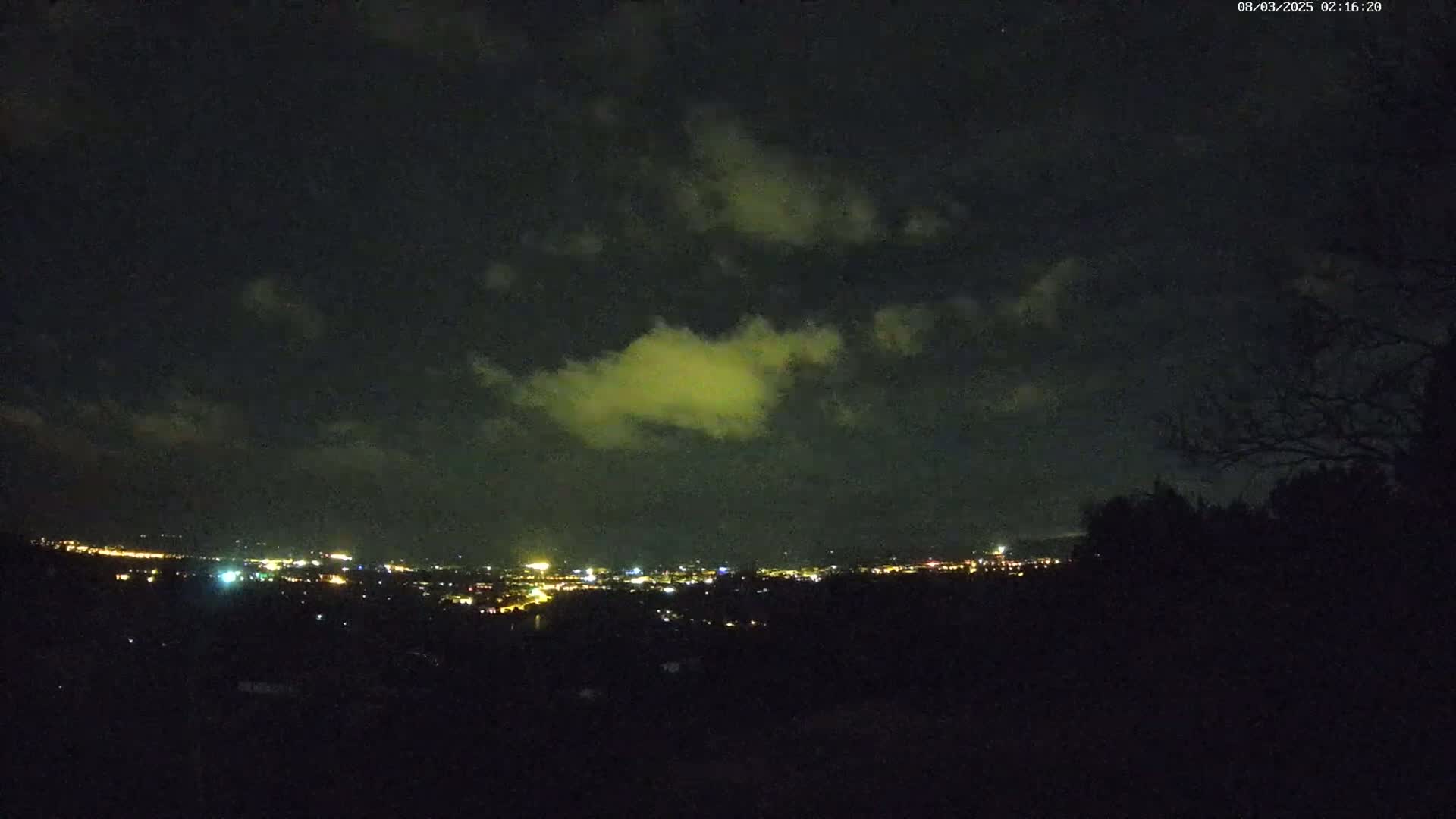 A nighttime view of a city's lights spread across a valley under a partly cloudy sky.